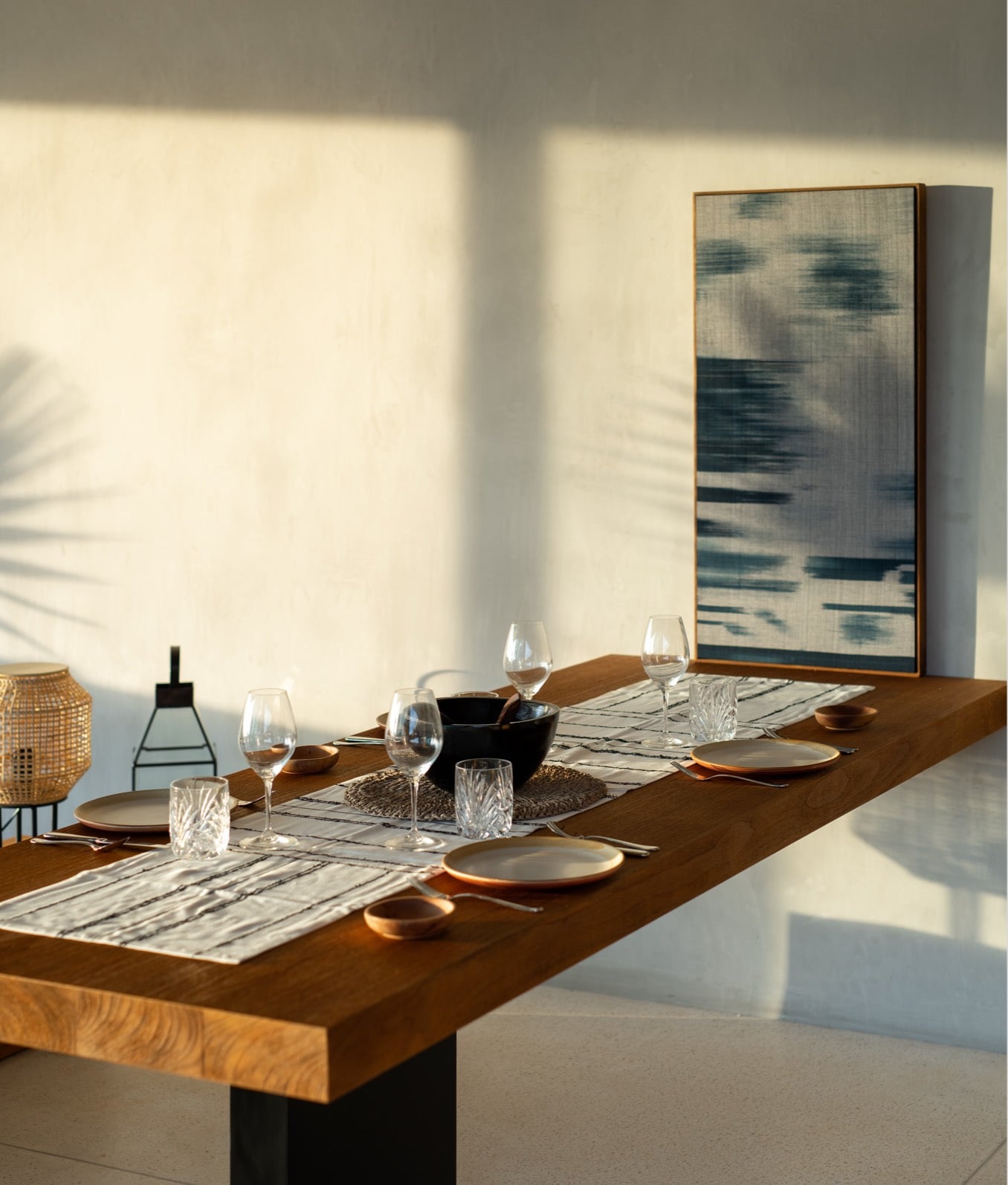 Cap Karoso, Asia — Wooden dining table set with wine glasses, ceramic plates, and natural light casting geometric shadows on white walls, contemporary art above.