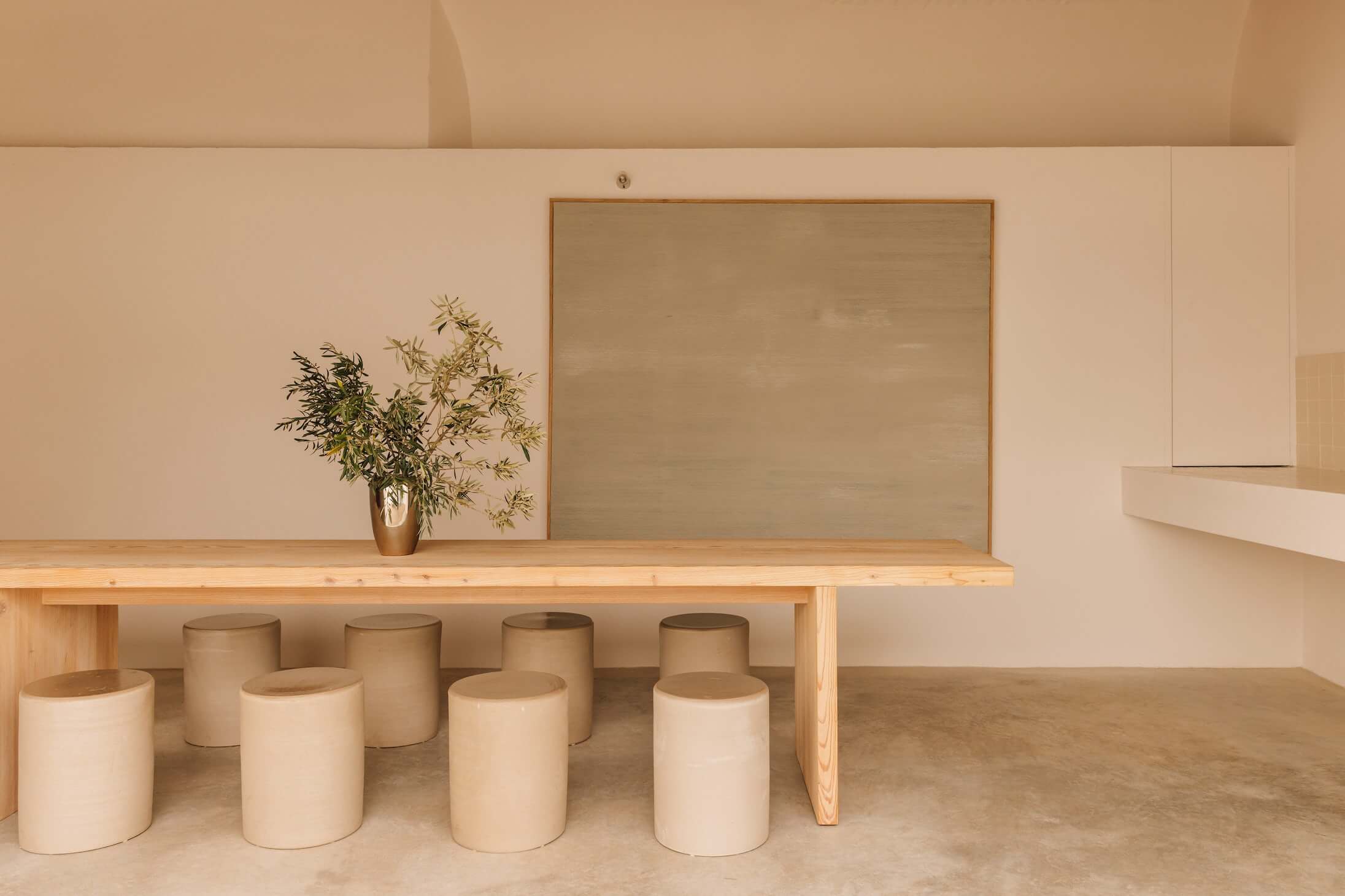 Casa Quatro, Europe — Minimalist dining space with natural wood table, cylindrical seating stools, framed artwork, potted plant, and warm neutral palette.