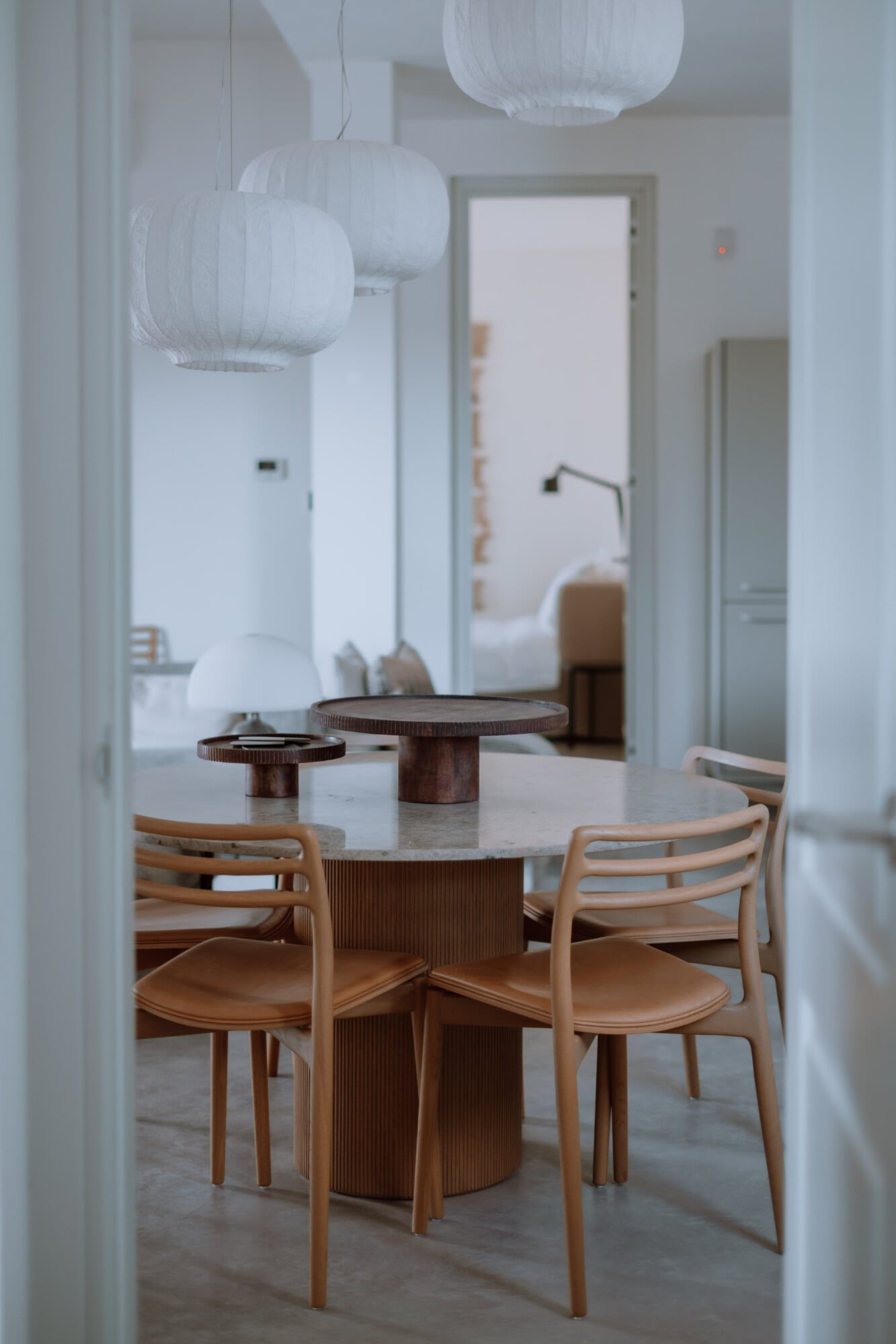 Villa Quercia, Italy — Minimalist dining room with round wood table, natural leather chairs, white paper pendant lights, and soft natural daylight filtering through open doorway.