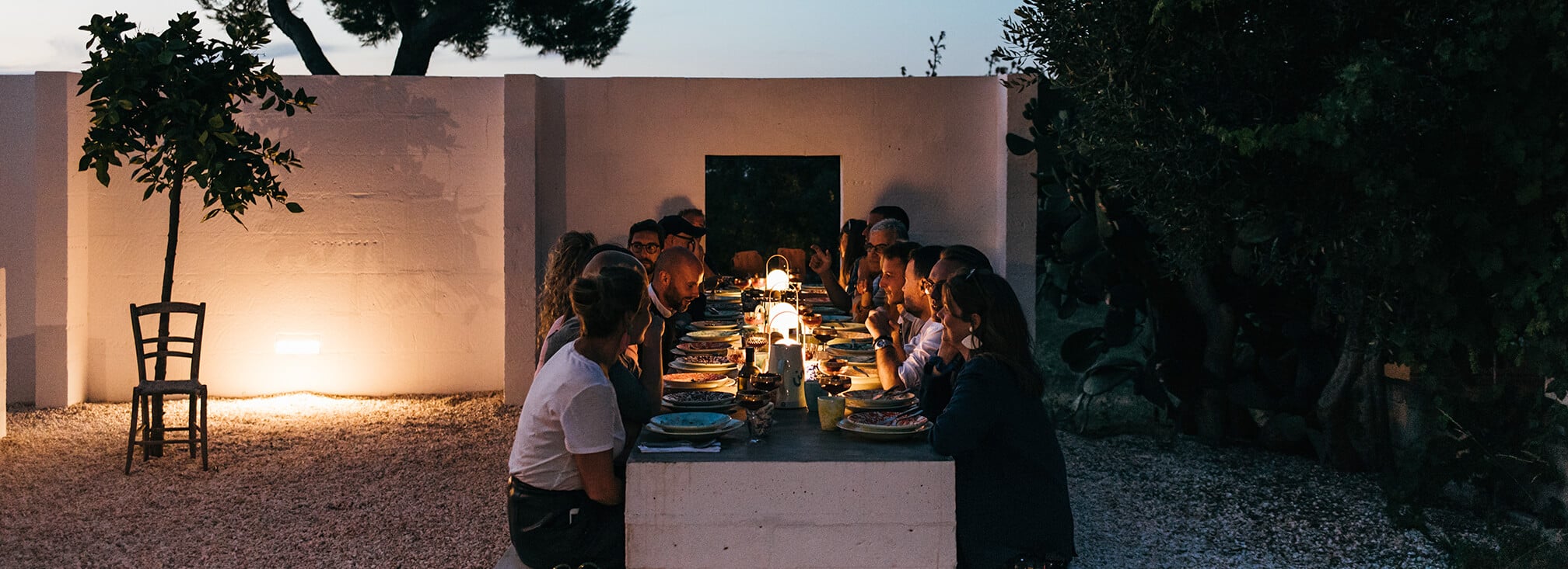 Masseria Moroseta, Europe — Long outdoor dining table illuminated by lanterns at dusk, white-walled courtyard with olive trees and ambient evening light.