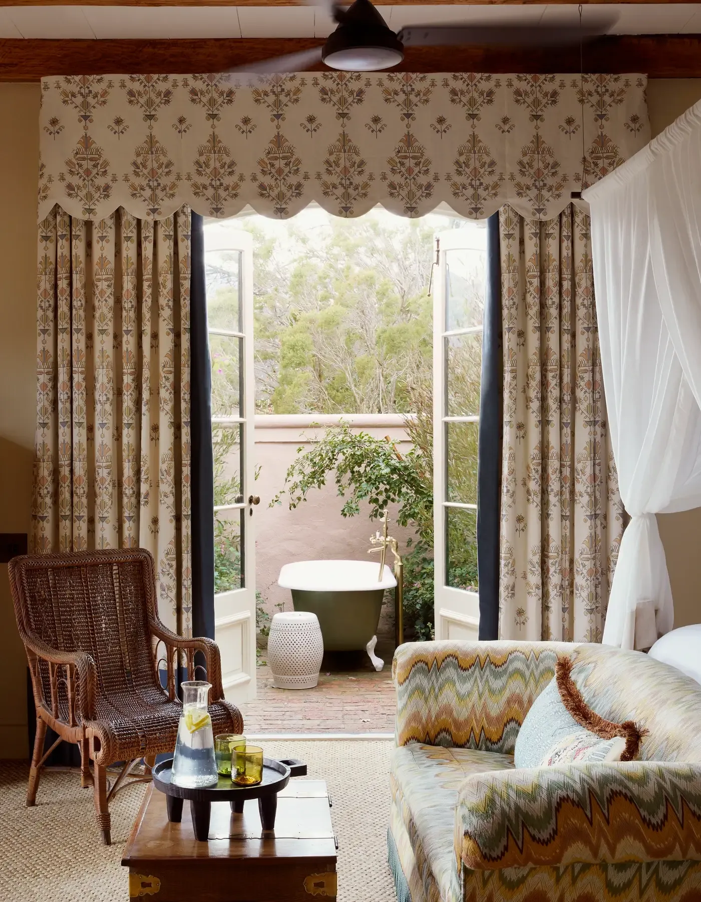 Sterrekopje, Africa — Luxury bedroom suite with damask curtains, wicker chair, patterned sofa, and glass doors opening to outdoor bathtub and landscape views.