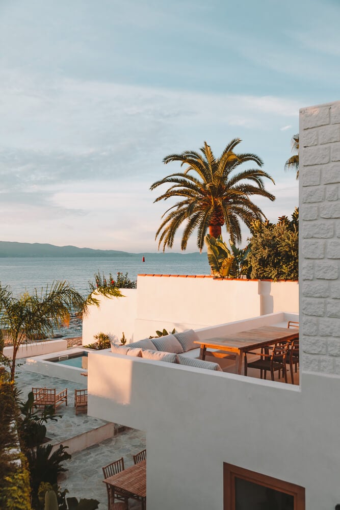 Casa Santa Teresa, Europe — Whitewashed Mediterranean villa terrace with palm tree, sea view, infinity pool, wooden dining furniture, and golden hour light casting long shadows.