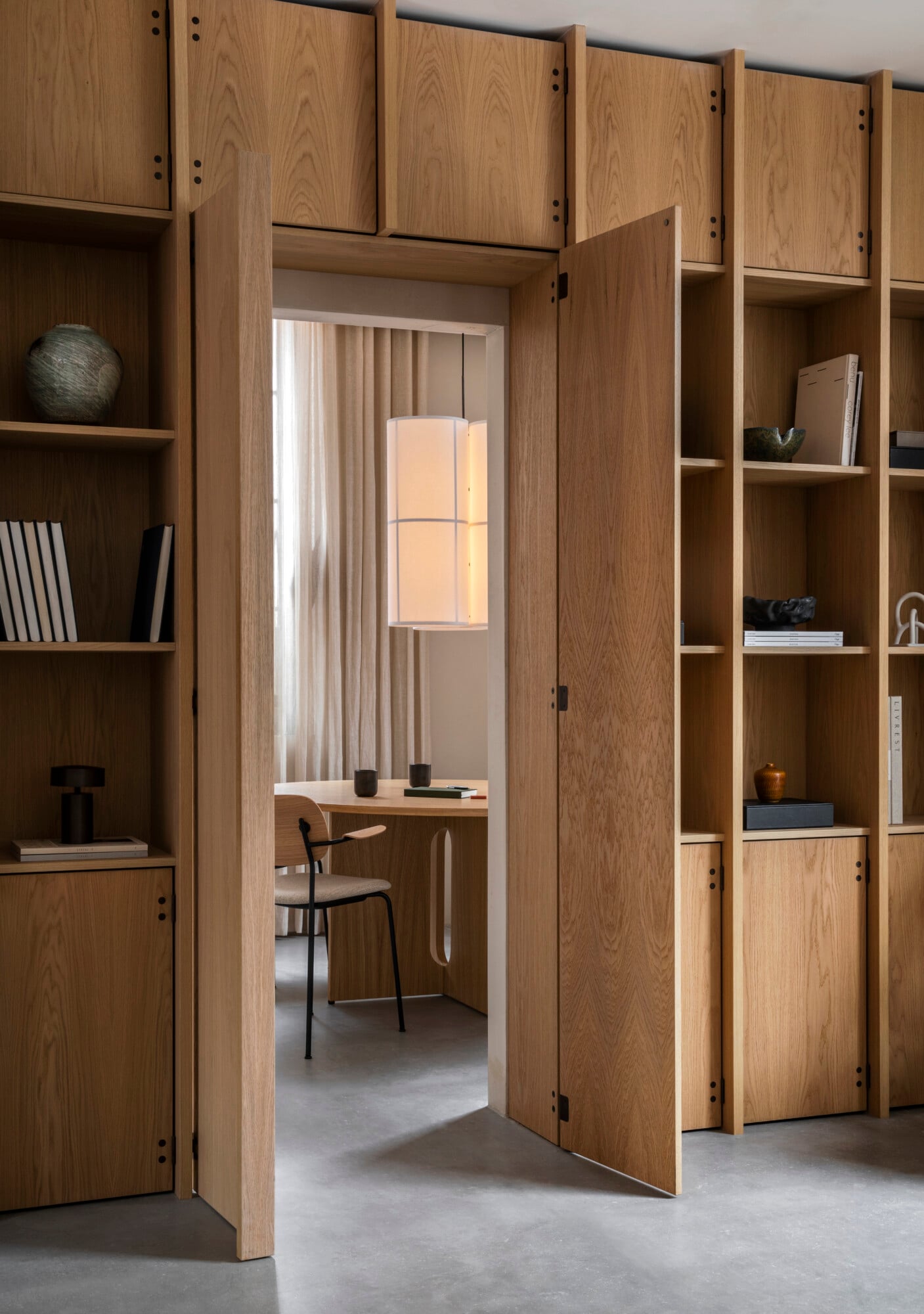 The Audo, Europe — Warm oak cabinetry with open shelving frames a pivoting wooden door to a minimalist workspace, pendant lighting casting soft glow on polished concrete floors.