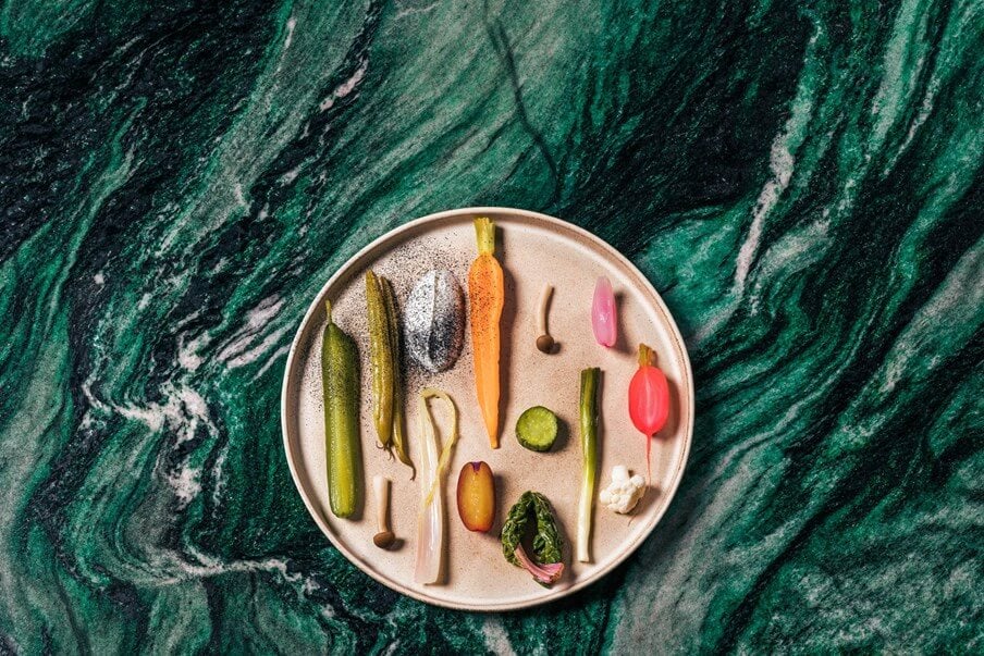 The Cōmodo — Cōmodo, Europe — Flat-lay of sliced vegetables and herbs arranged on cream plate against green marble surface, editorial food styling.