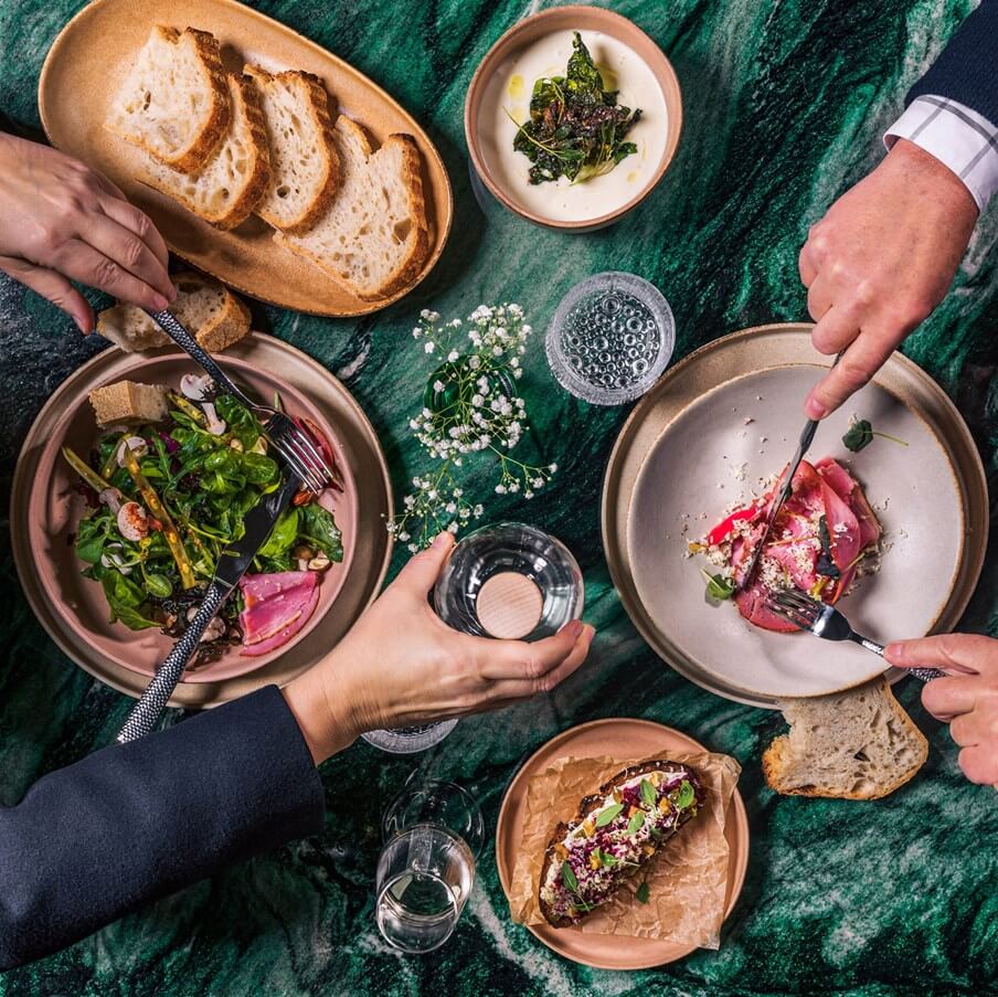 the cōmodo restaurant — Cōmodo Restaurant, Europe — Overhead view of plated dishes with beetroot, leafy greens, bread, and hands dining at marble table in jewel-toned setting.