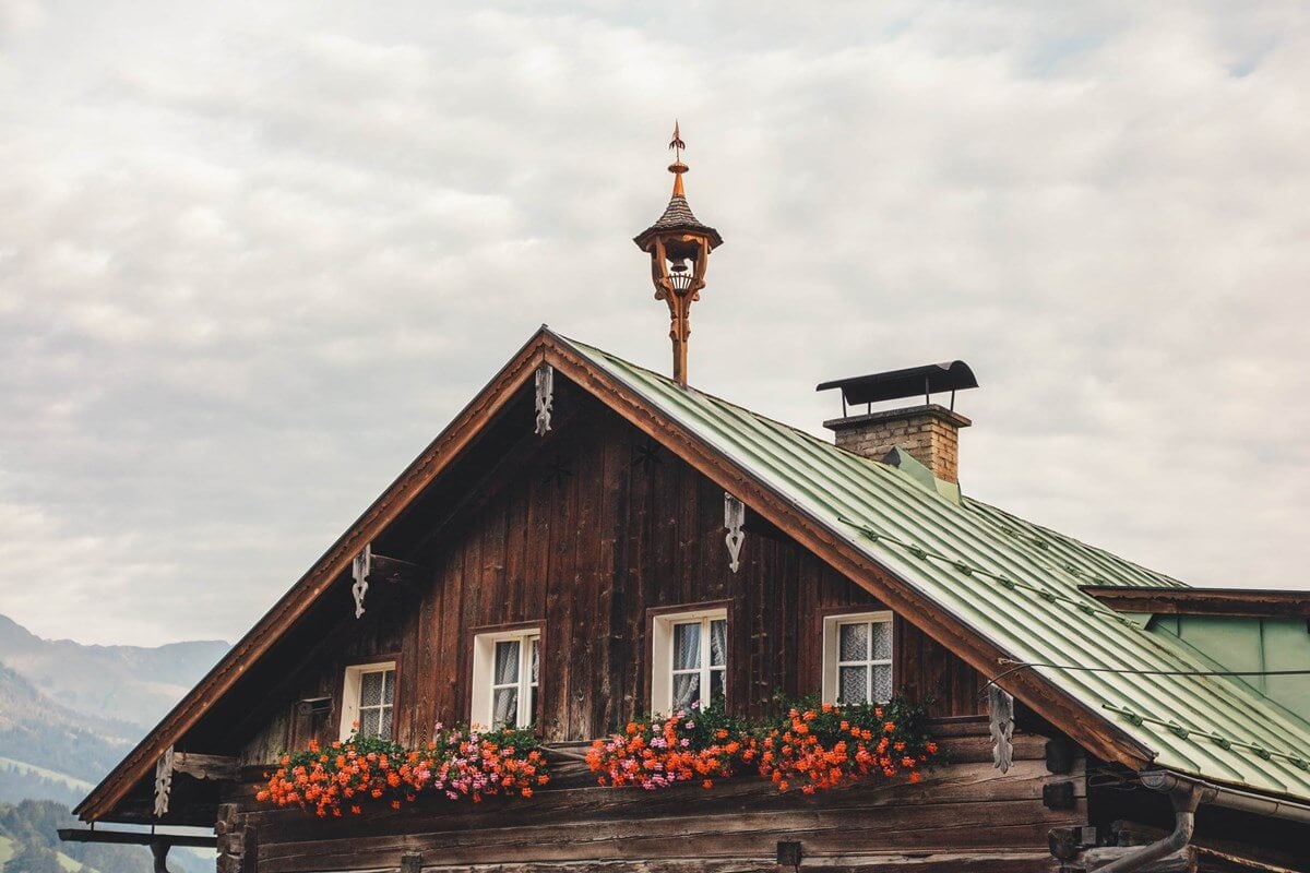 The Cōmodo — Cōmodo, Europe — Dark wood alpine chalet with green metal roof, ornamental carved details, bright orange geraniums in window boxes, mountain valley backdrop.