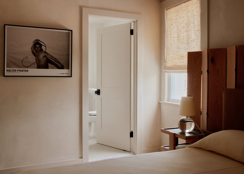 The James Bradley, North America — Minimalist bedroom with warm neutral tones, open bathroom, wooden accents, natural light, and contemporary art.