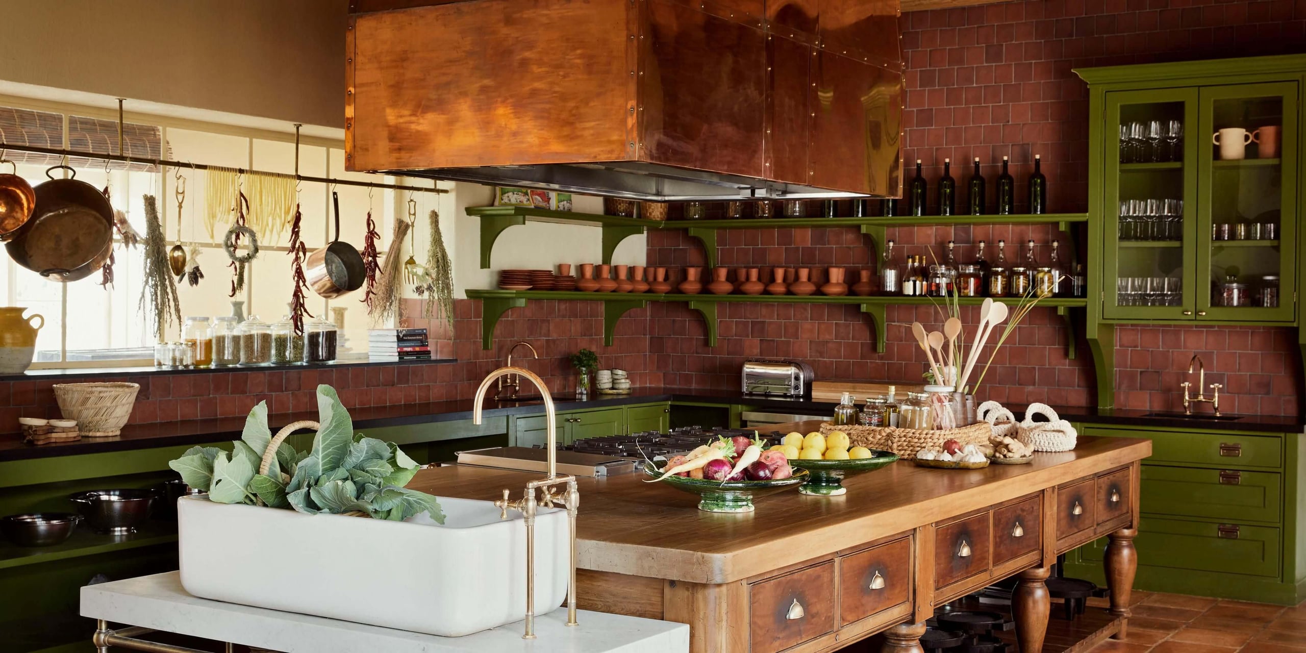 Sterrekopje, Africa — Terracotta-tiled kitchen with sage-green cabinetry, copper hood, wooden island, fresh produce, and hanging herbs by natural light.