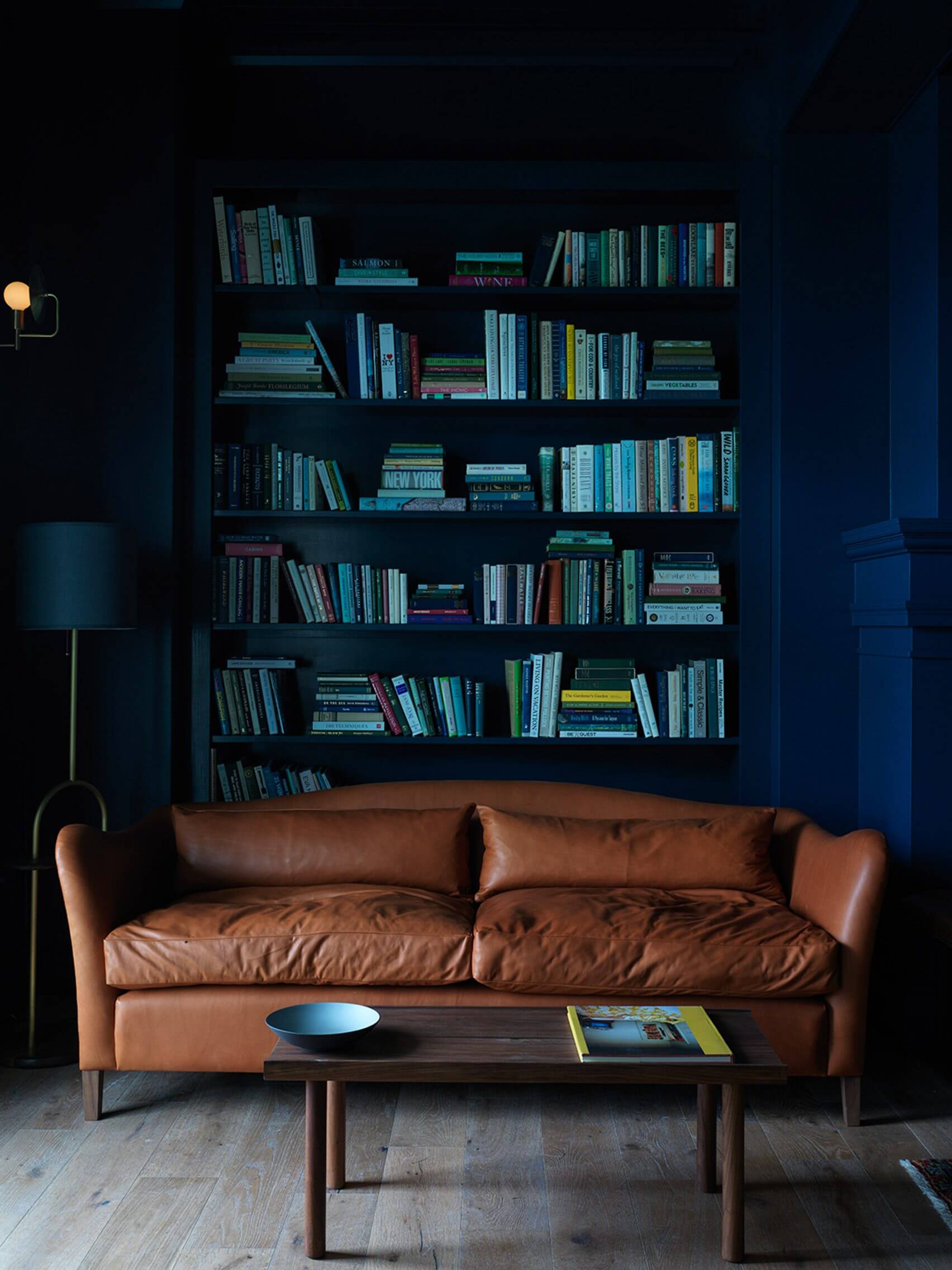 The Lake House, North America — Cognac leather sofa before dark blue shelving filled with books, mid-century modern interior with warm wood flooring and brass floor lamp.