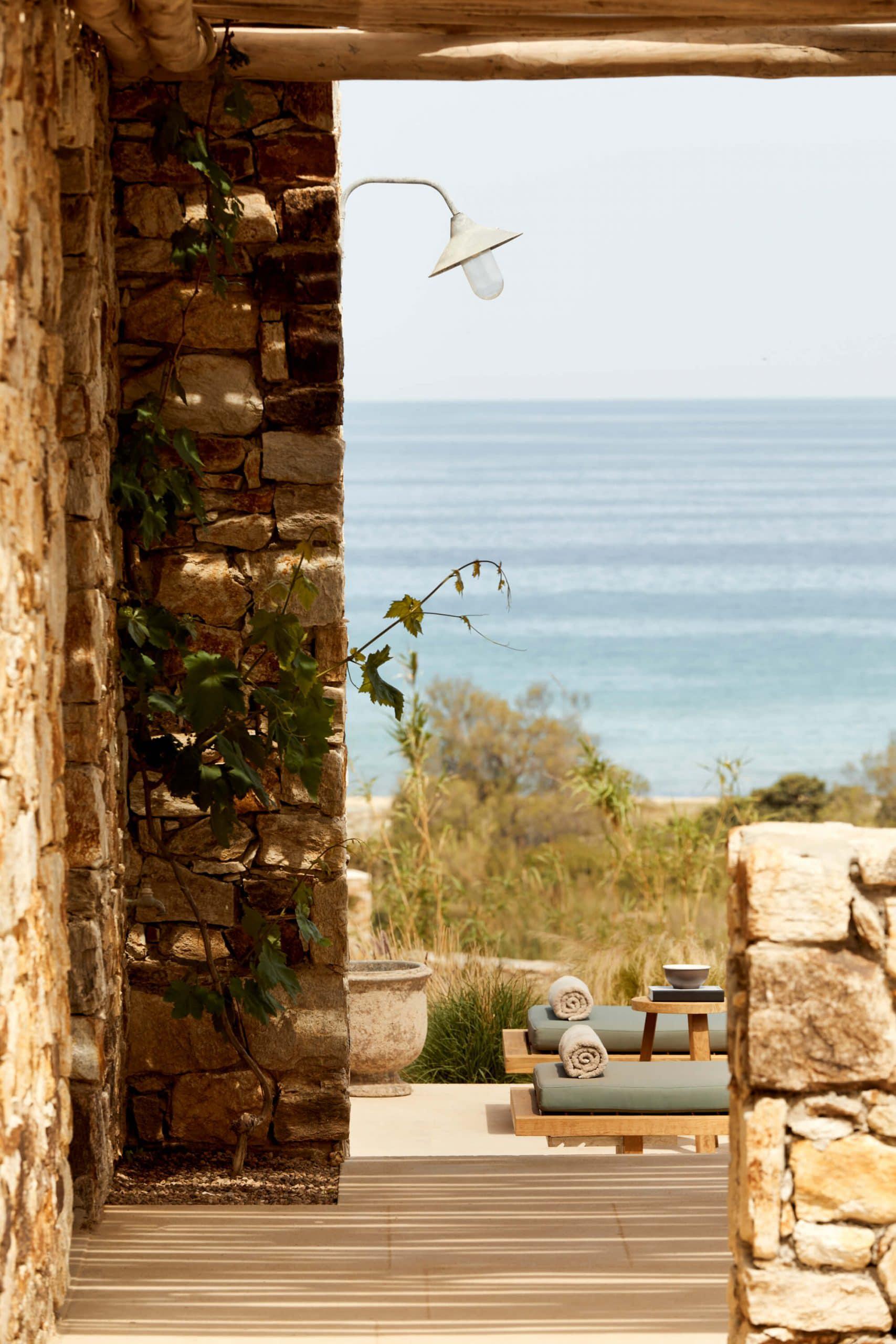 The Rooster, Europe — Stone archway framing coastal vista, wooden lounge chairs with rolled linens, vintage wall sconce, sea-view terrace with Mediterranean architecture.
