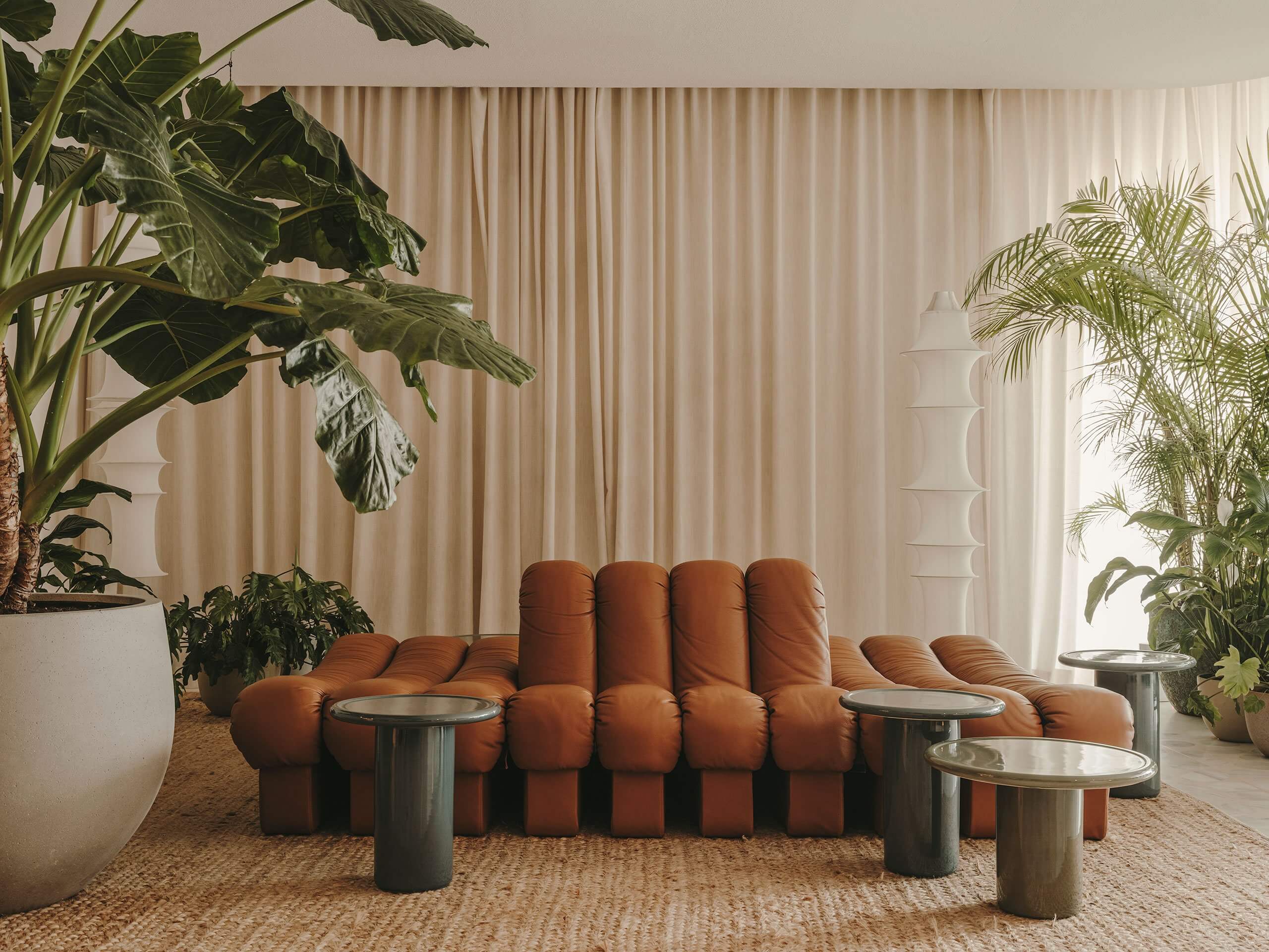 The Standard Ibiza, Balearic Islands, Spain — Terracotta puffy sofa with metallic side tables surrounded by potted tropical plants and cream curtains in minimalist lounge space.