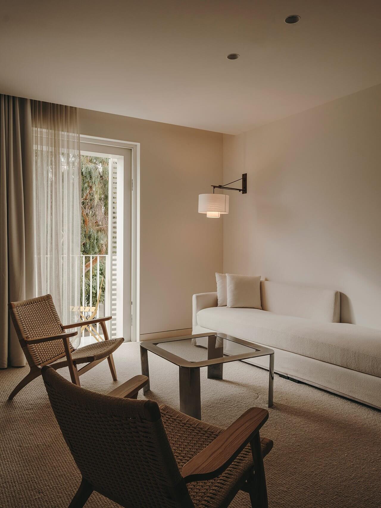 The Standard Ibiza, Balearic Islands, Spain — Mid-century modern living room with woven lounge chairs, cream sofa, glass coffee table, and sheer curtains framing a sunlit balcony view.
