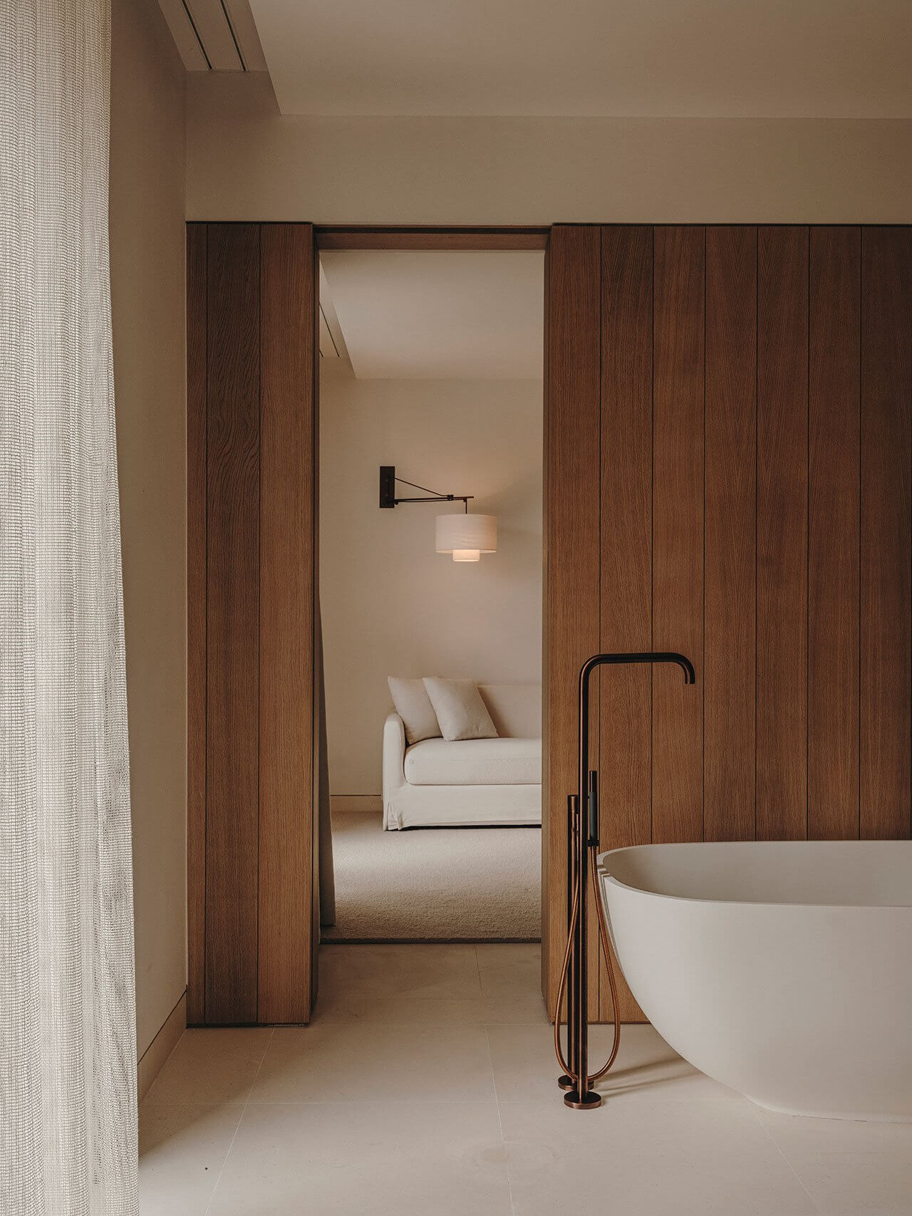 The Standard Ibiza — Standard Ibiza, Balearic Islands, Spain — Minimalist bathroom with freestanding tub, dark wood partition doors opening to bedroom with cream sofa, pendant lighting, neutral palette.