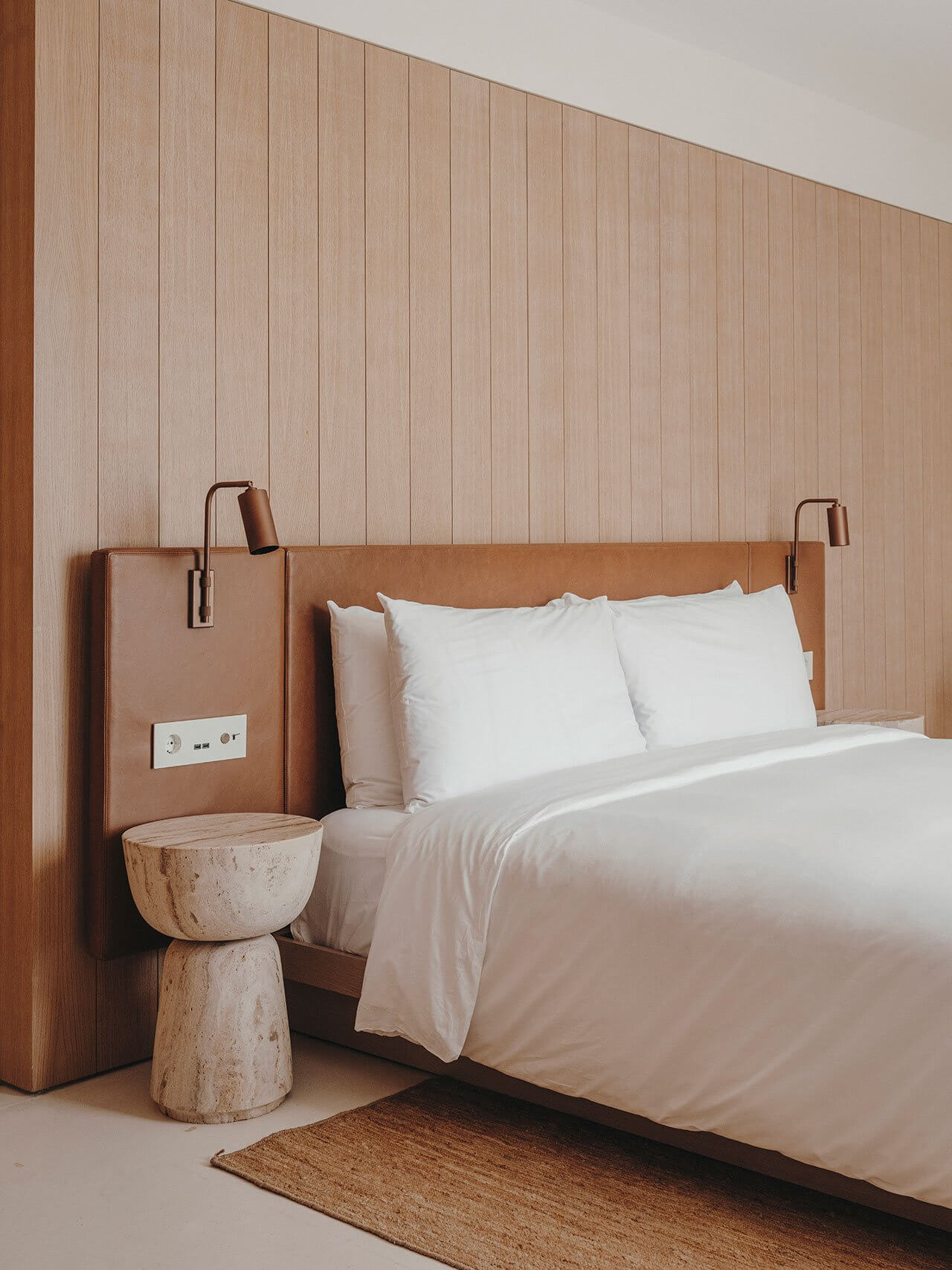 The Standard Ibiza, Balearic Islands, Spain — Minimalist hotel bedroom with cognac leather headboard, white linens, travertine side table, and vertical wood-paneled wall.