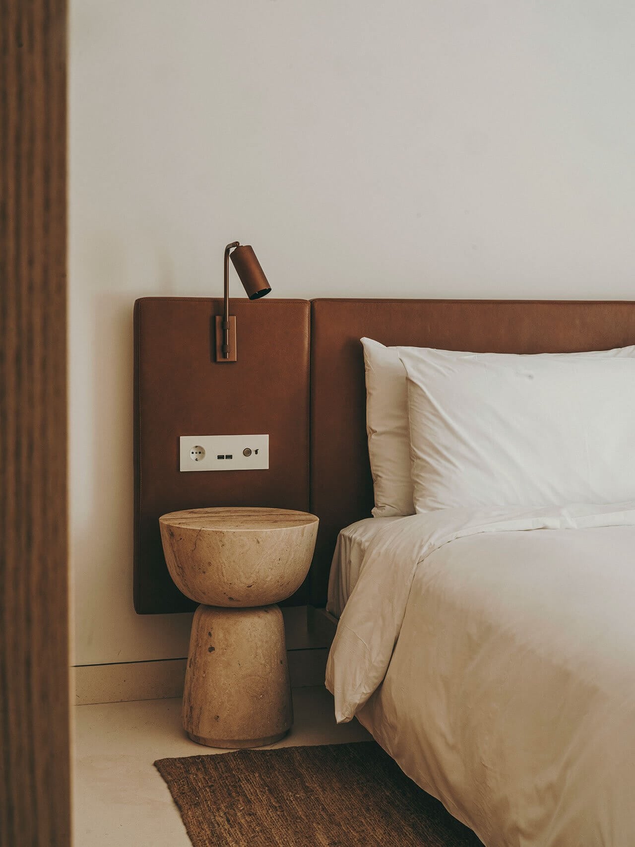The Standard Ibiza, Balearic Islands, Spain — Minimalist bedroom with cognac leather headboard, wooden pedestal side table, brass reading light, white linens, and neutral palette.