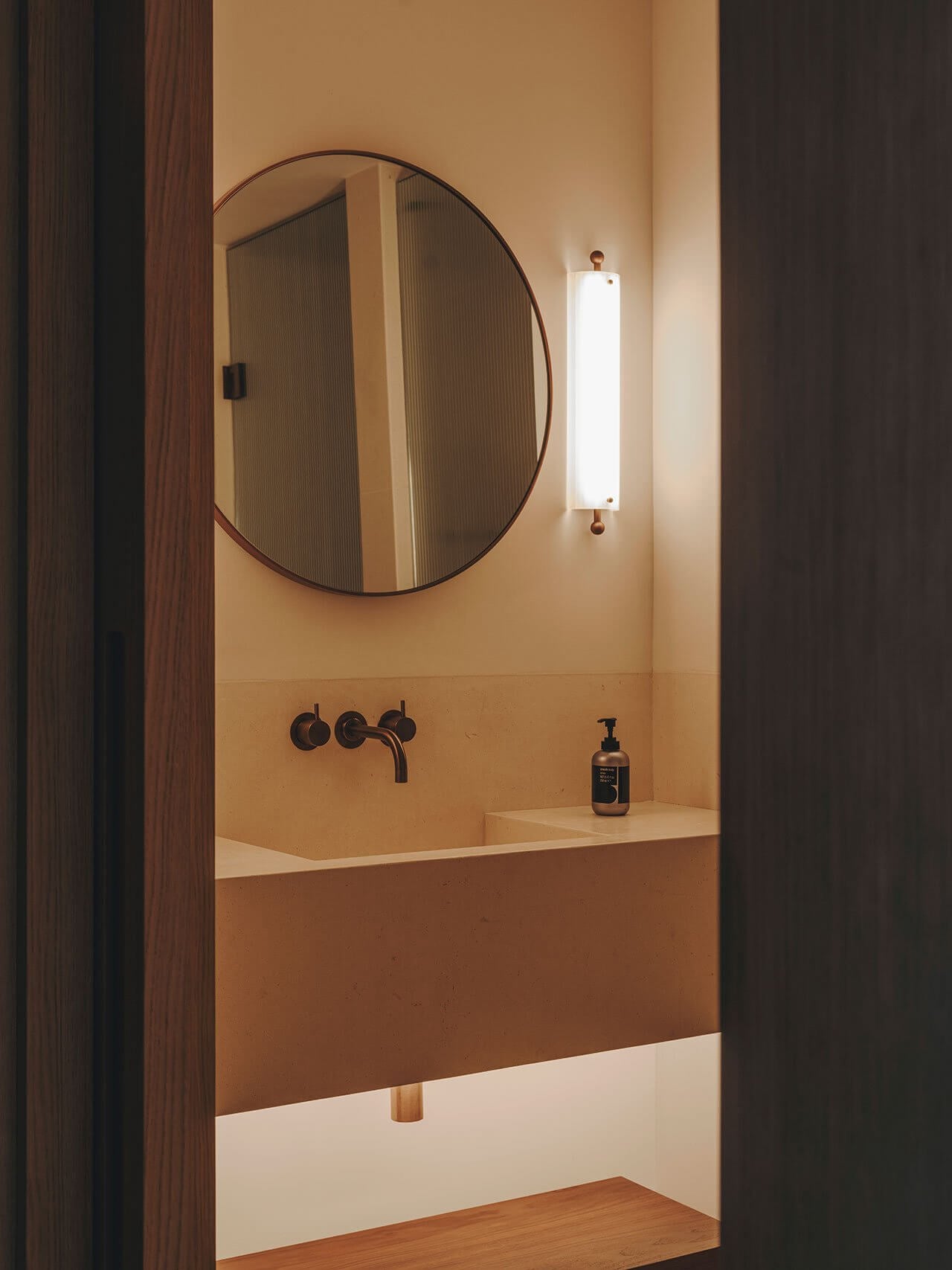 The Standard Ibiza, Balearic Islands — Minimalist bathroom with circular mirror, terracotta walls, black fixtures, and vertical sconce lighting above concrete sink.