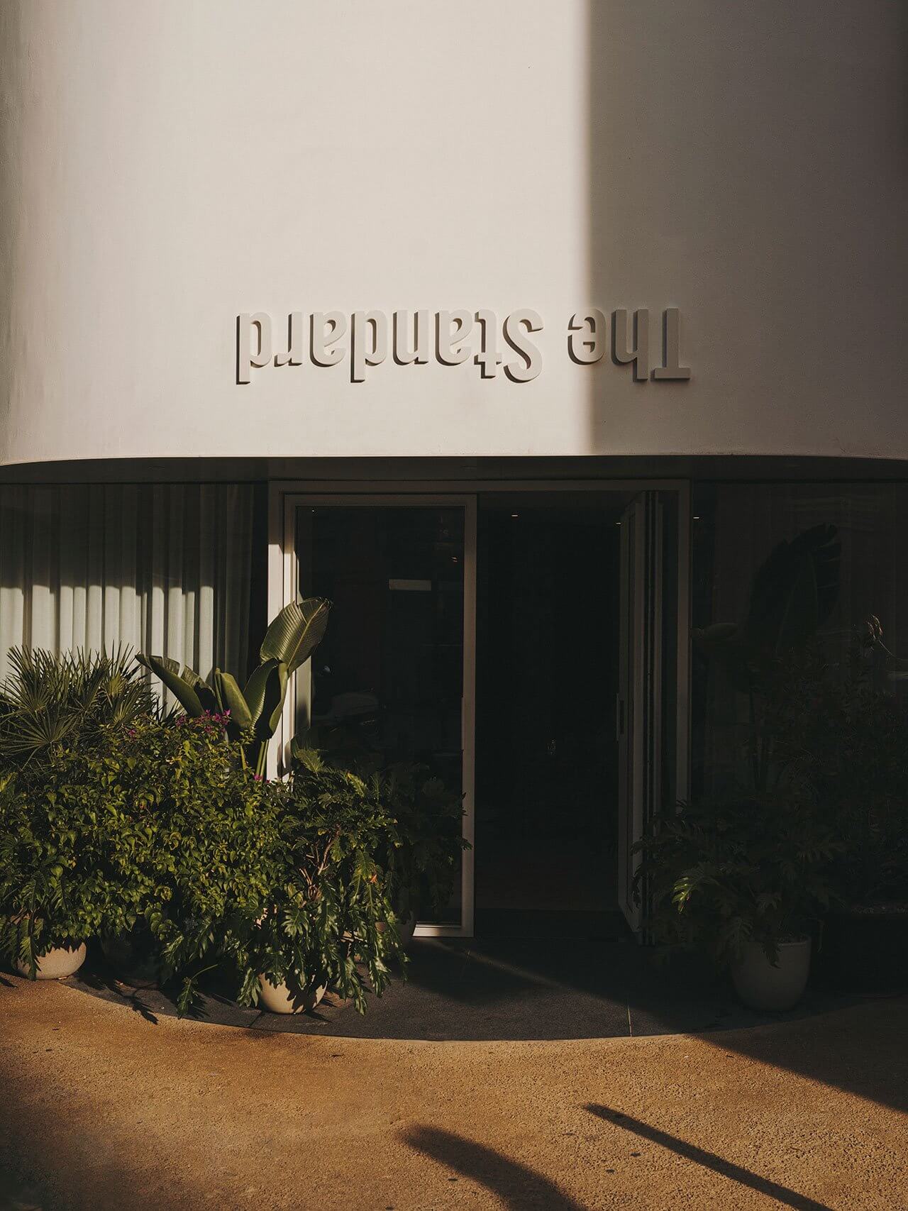 The Standard Ibiza, Balearic Islands, Spain — Minimalist entrance with white facade, dark glazed doors, potted plants, and geometric shadows cast by afternoon sunlight.