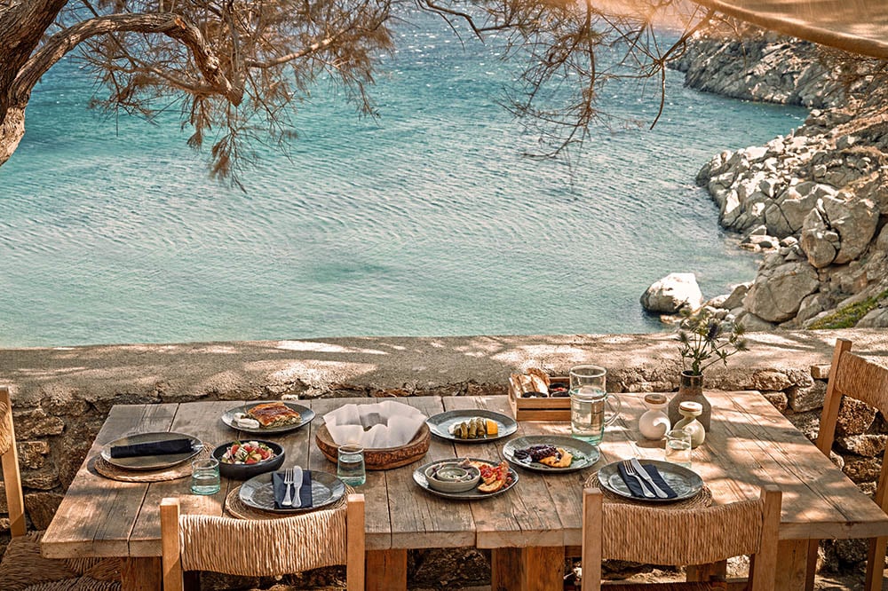 The Wild, Europe — Seaside dining table set with Mediterranean plates and glassware beneath pine branches, turquoise water and rocky coastline beyond.
