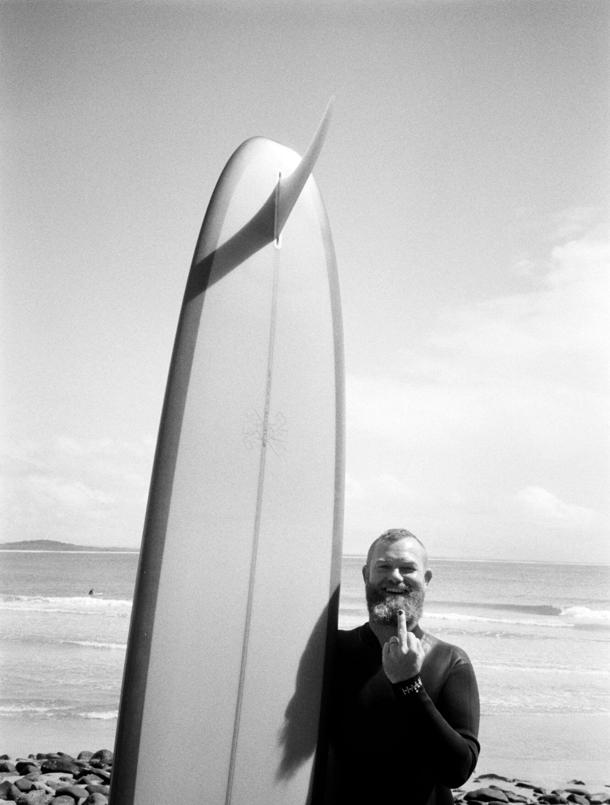 Thomas + Smile, Asia-Pacific — Bearded man with surfboard on rocky beach at dawn, minimalist coastal aesthetic, black wetsuit, serene ocean landscape.