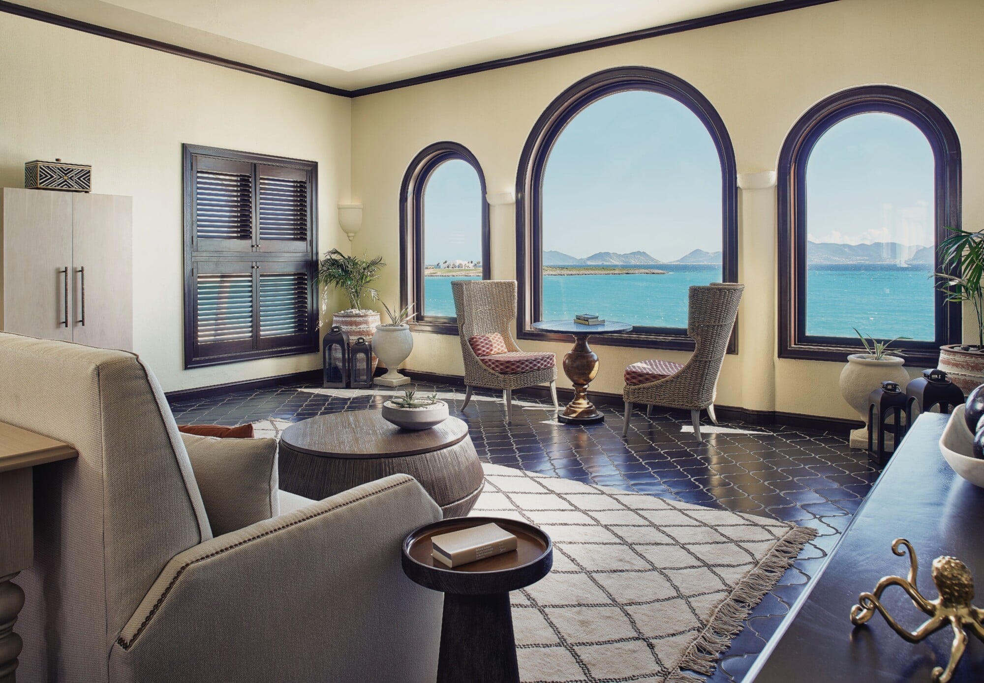 Cap Juluca, North America — Coastal suite with arched windows overlooking turquoise bay, woven chairs, cream walls, dark tile flooring, modern minimalist furnishings.