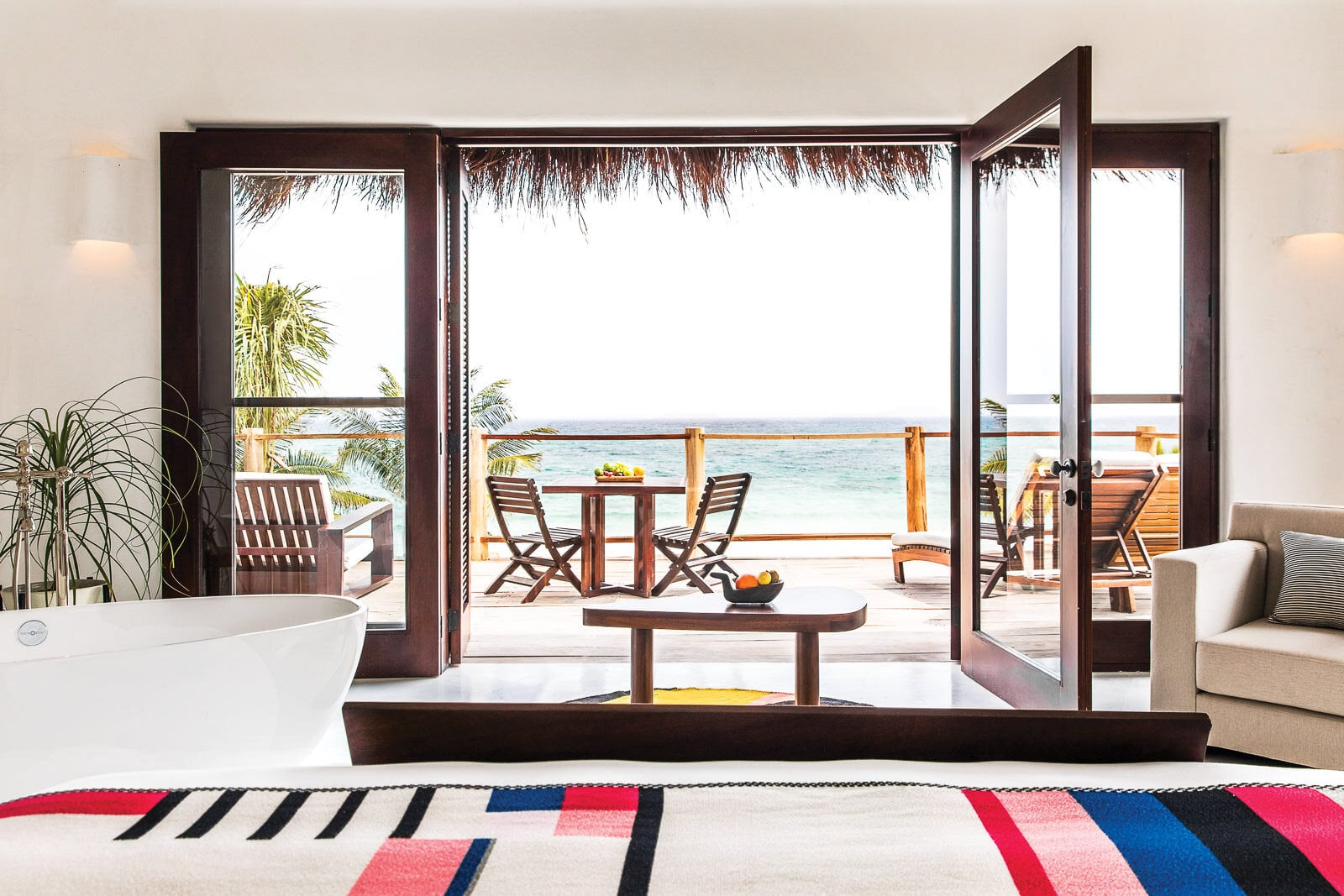 Hotel Esencia, Mexico — Oceanfront suite with dark wood doors opening to beachside deck, minimalist white bathtub, natural light, tropical palm views, contemporary luxury resort design.