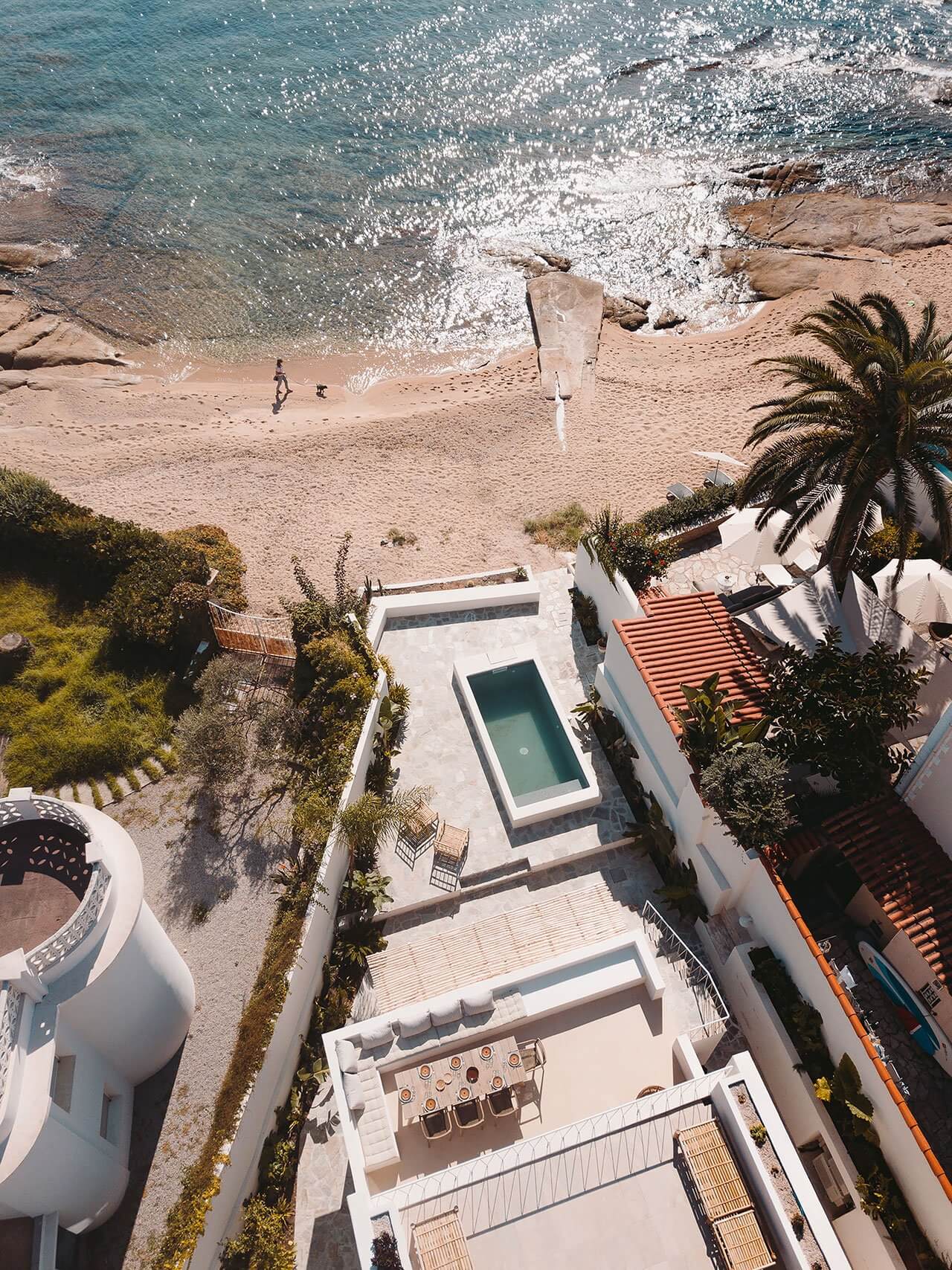 Casa Santa Teresa, Europe — Aerial view of modernist beachfront villa with terracotta roof, infinity pool, and Mediterranean coast with turquoise waters and sandy beach.