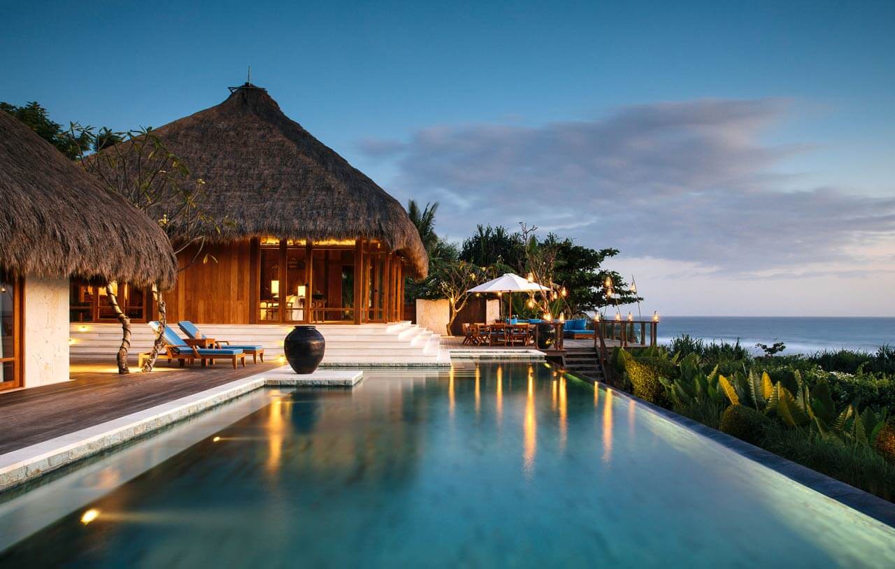NIHI Sumba, Indonesia — Beachfront villa with thatched roof, infinity pool at dusk, wooden architecture overlooking ocean, tropical landscape lighting.