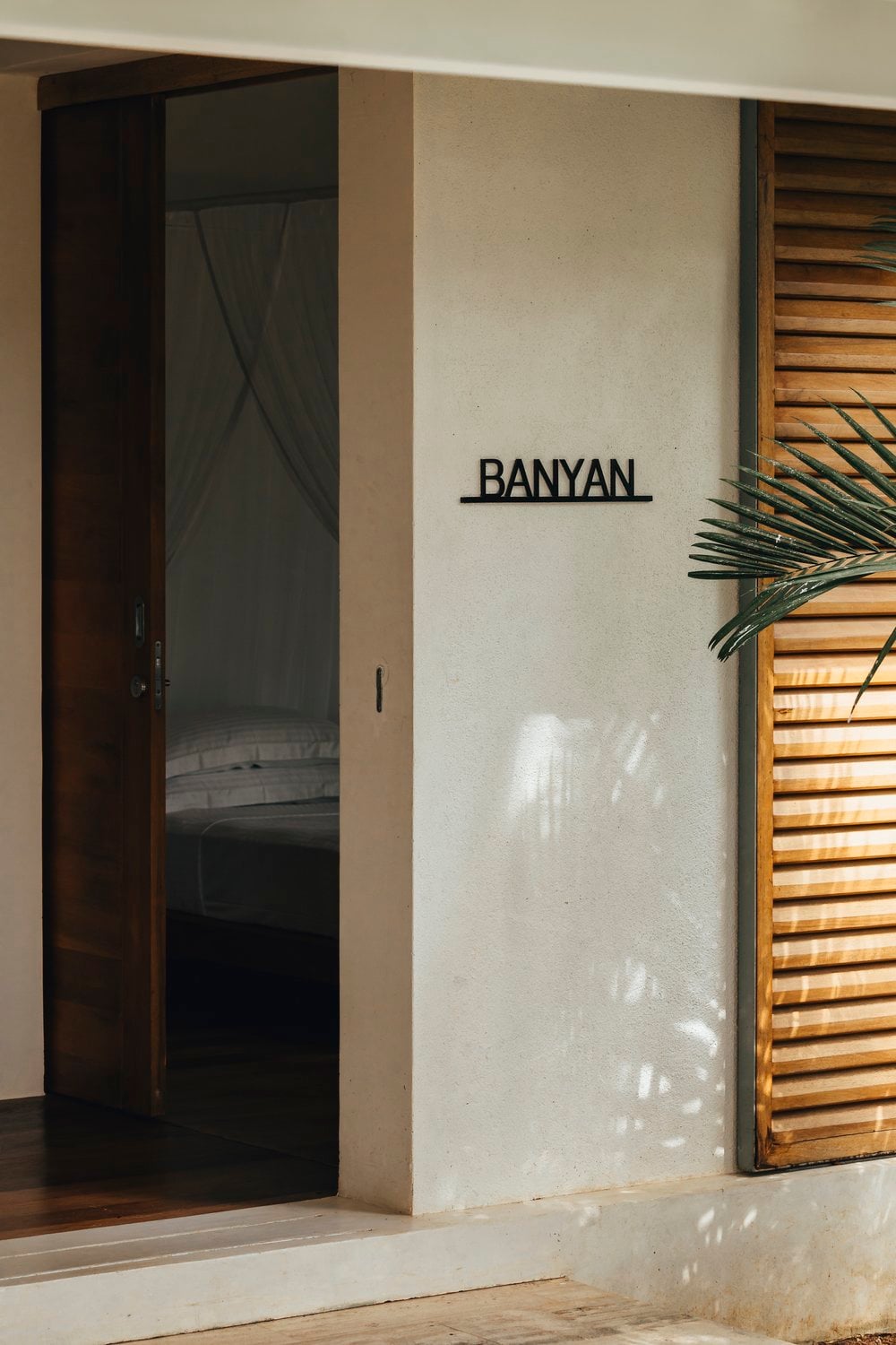 Kurulu Bay, Asia — Minimalist villa entrance with dark timber door, cream concrete walls, wooden louvered shutters, and tropical plant, morning light casting geometric shadows.