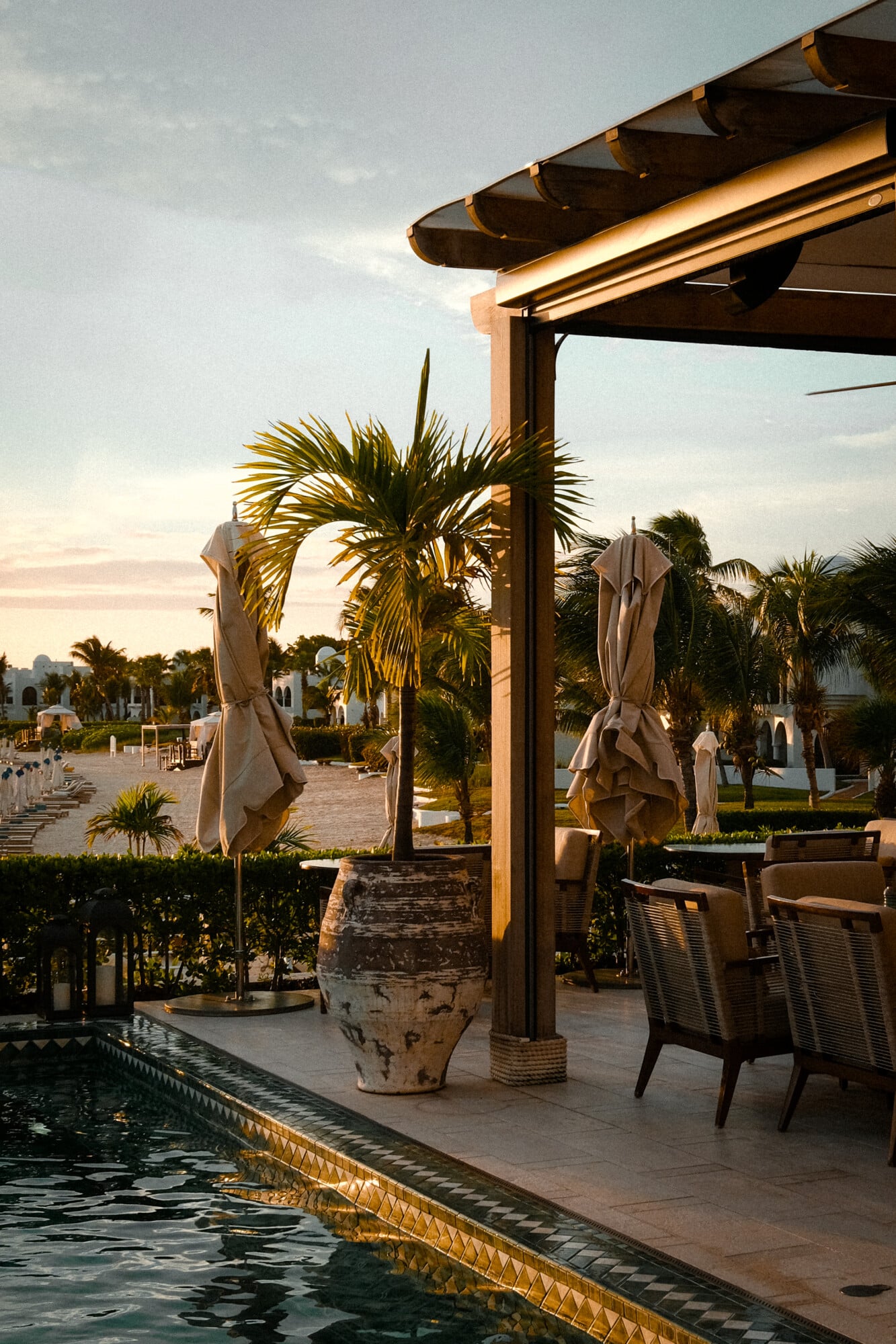 Uchu, North America — Pergola-shaded terrace with draped umbrellas, palm plants, and poolside seating at golden hour, Mediterranean-inspired luxury hospitality design.