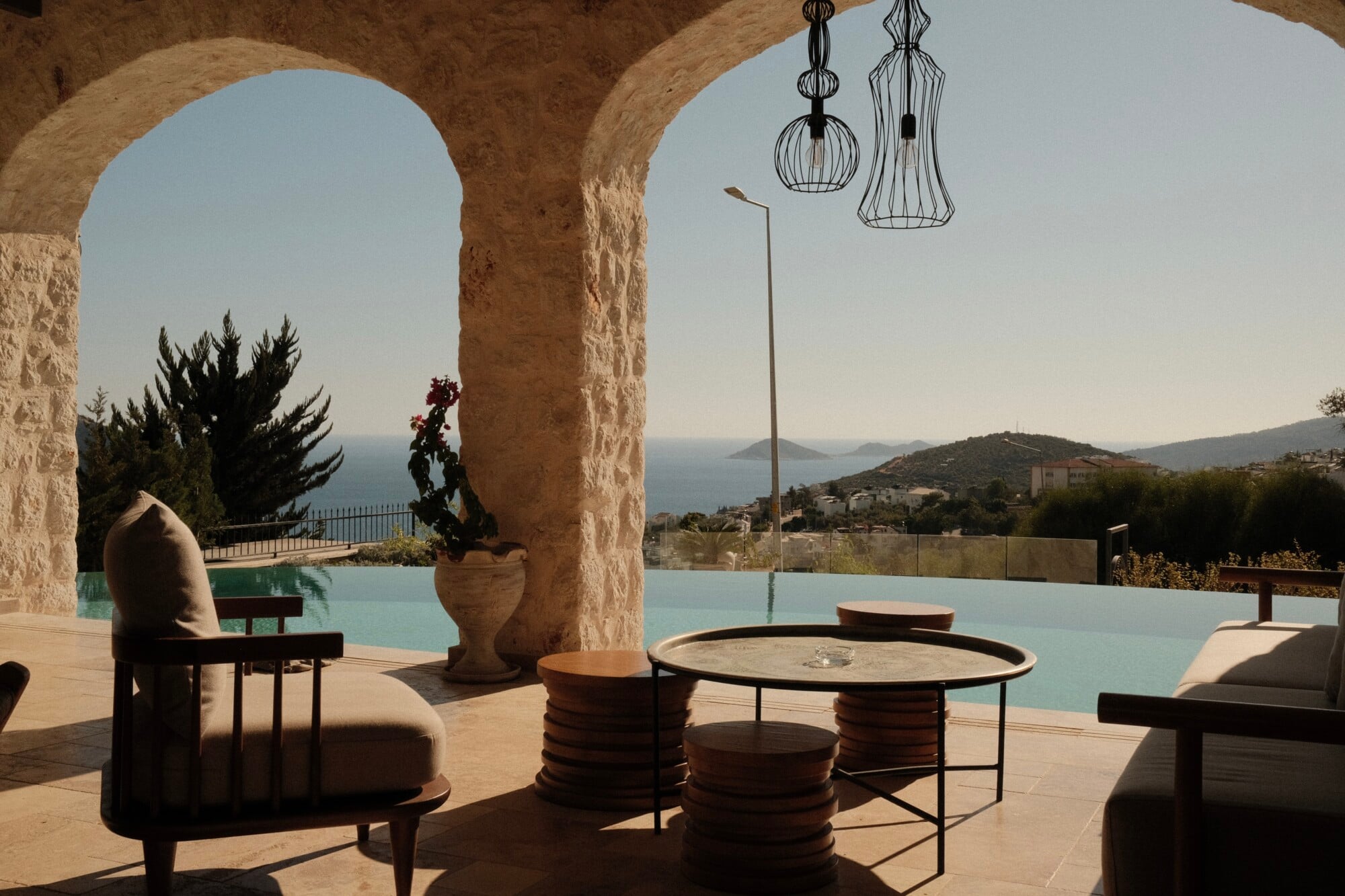 Villa Thalia by Muses Kalkan, Kalkan, Turkey — Stone archways frame turquoise pool overlooking Mediterranean coastline, minimalist wooden furniture and hanging pendant lights beneath travertine columns.