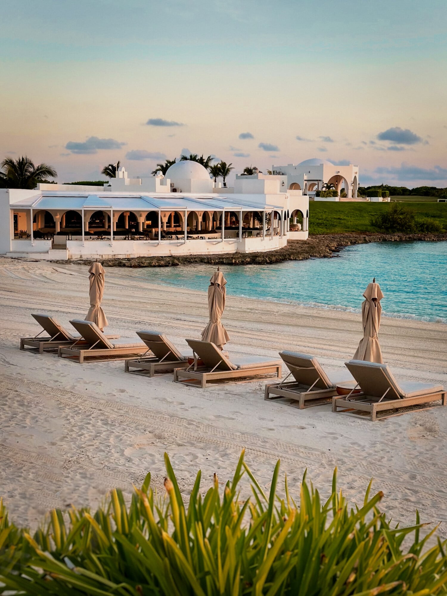 Cap Juluca, Anguilla — Whitewashed beachfront pavilion with arched colonnade, teak loungers, closed umbrellas, turquoise Caribbean waters at dusk.