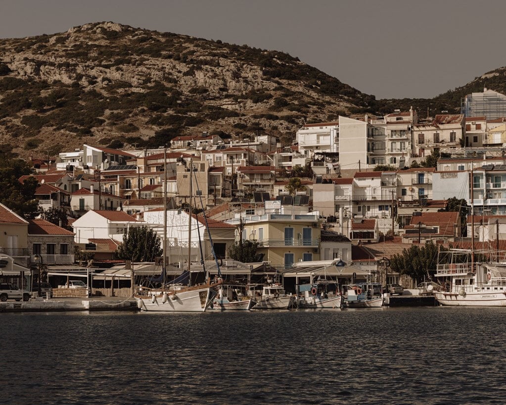 Village of Pythagorion — Waterfront village with terracotta-roofed buildings cascading up rocky hillside, traditional fishing boats moored at stone quay, Mediterranean coastal architecture and light.