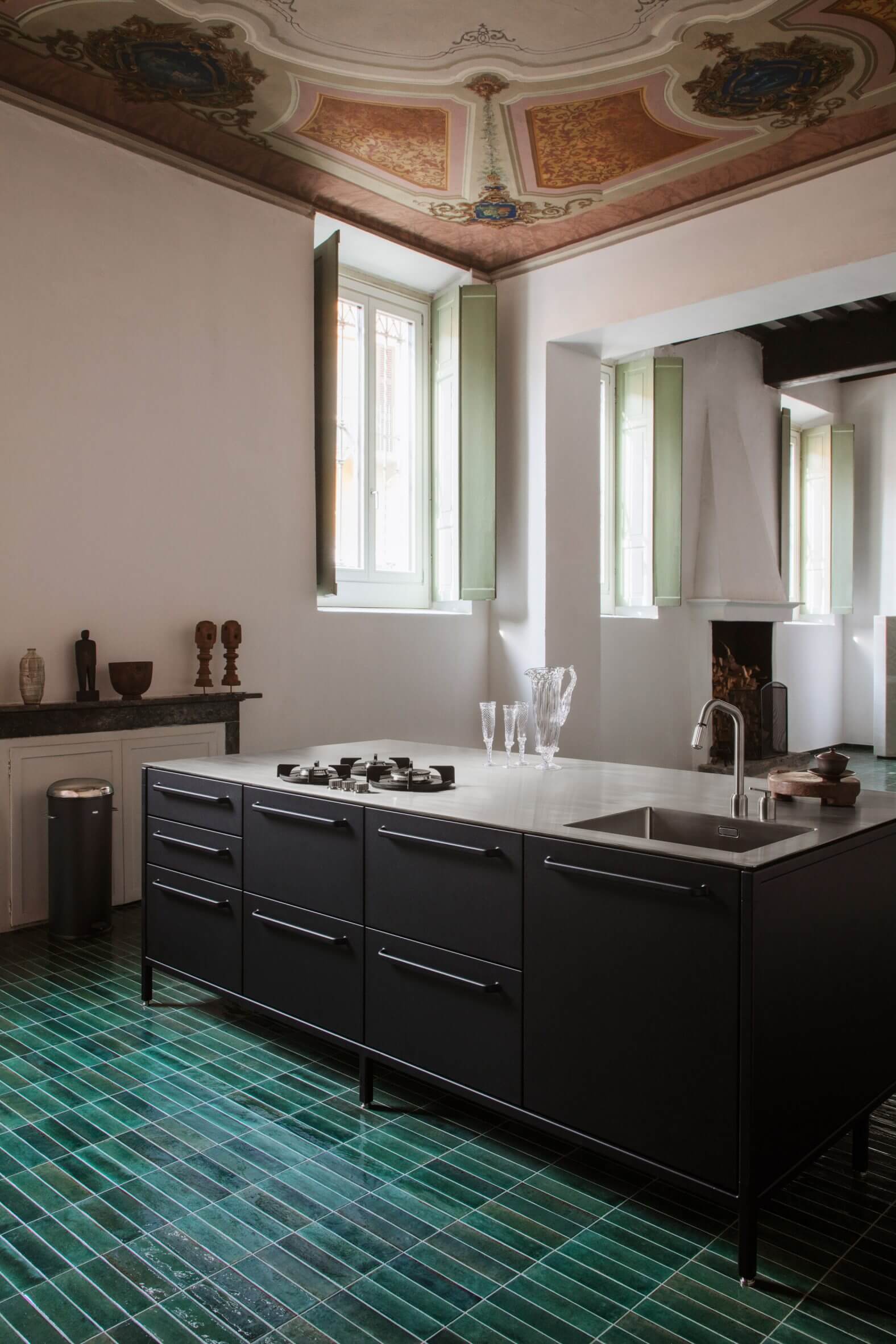 Vipp Palazzo Monti, Italy — Black kitchen island with white countertop, green tile floor, ornate frescoed ceiling, pale pink walls, modern luxury interior design.