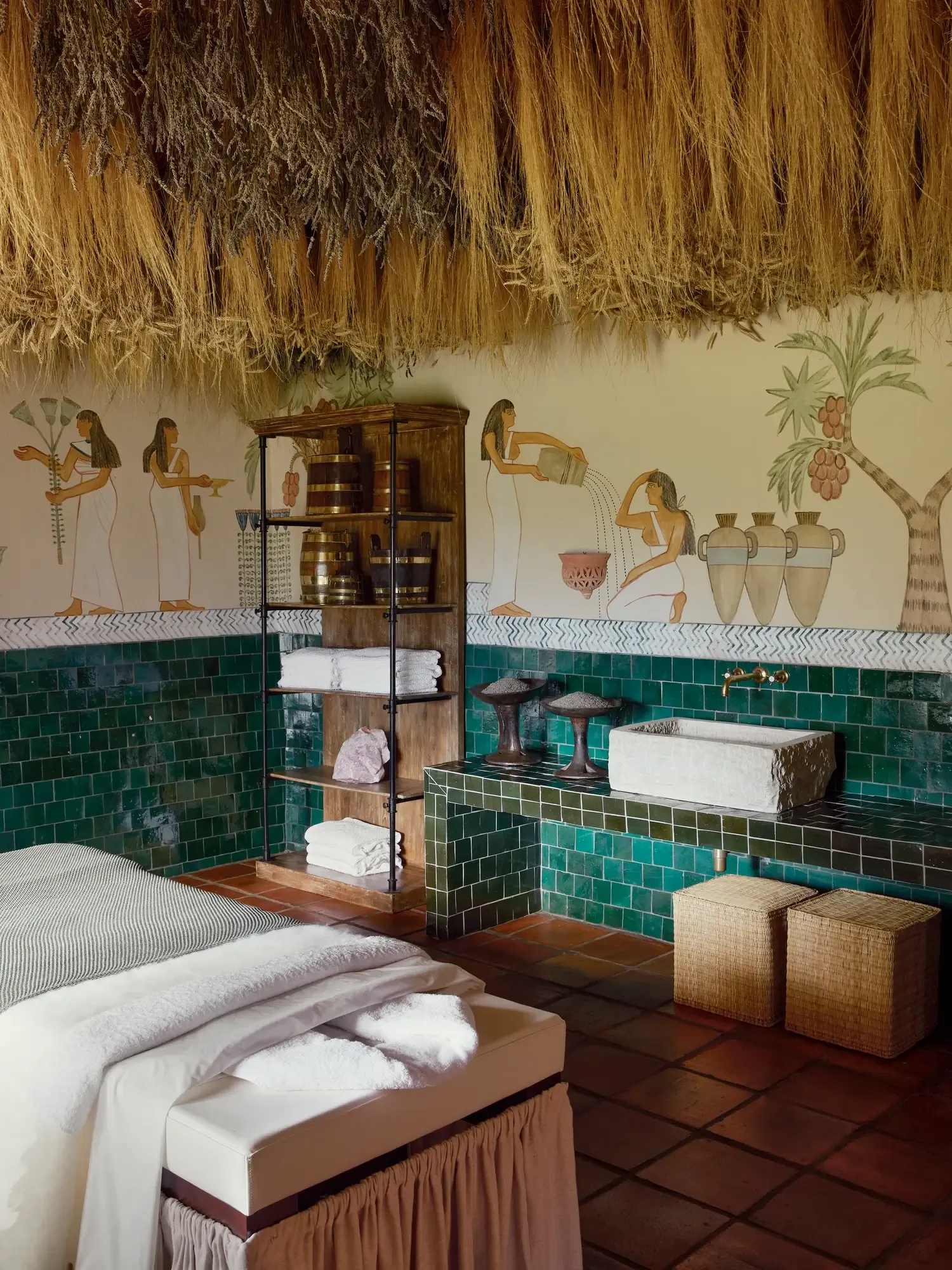 Sterrekopje, Africa — Egyptian-inspired bathroom with teal zellige tilework, terra-cotta floors, palm-thatch ceiling, and hieroglyphic murals above stone sink and soaking tub.