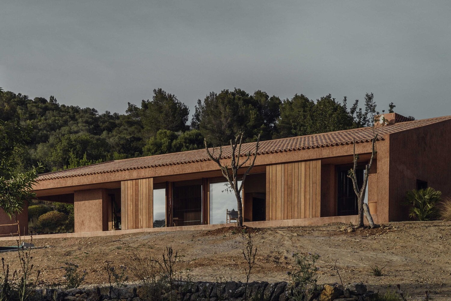 Alcúdia House, Balearic Islands — Terracotta-roofed residence with rammed-earth walls and wooden shutters overlooking Mediterranean landscape, minimalist contemporary design.