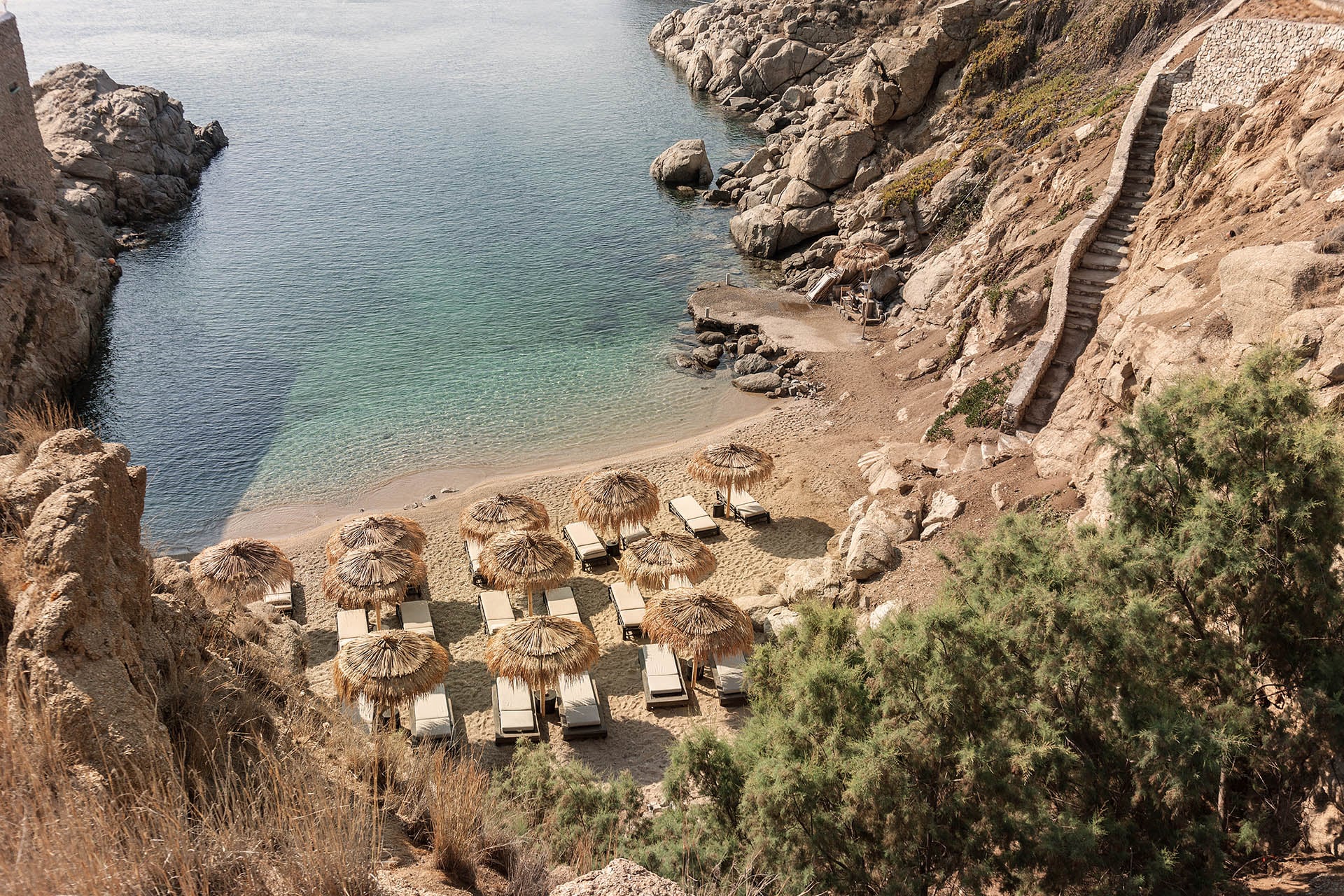 The Wild — Secluded Mediterranean beach cove with straw umbrellas, lounge chairs, turquoise water, dramatic rocky cliffs, and stone staircase access.