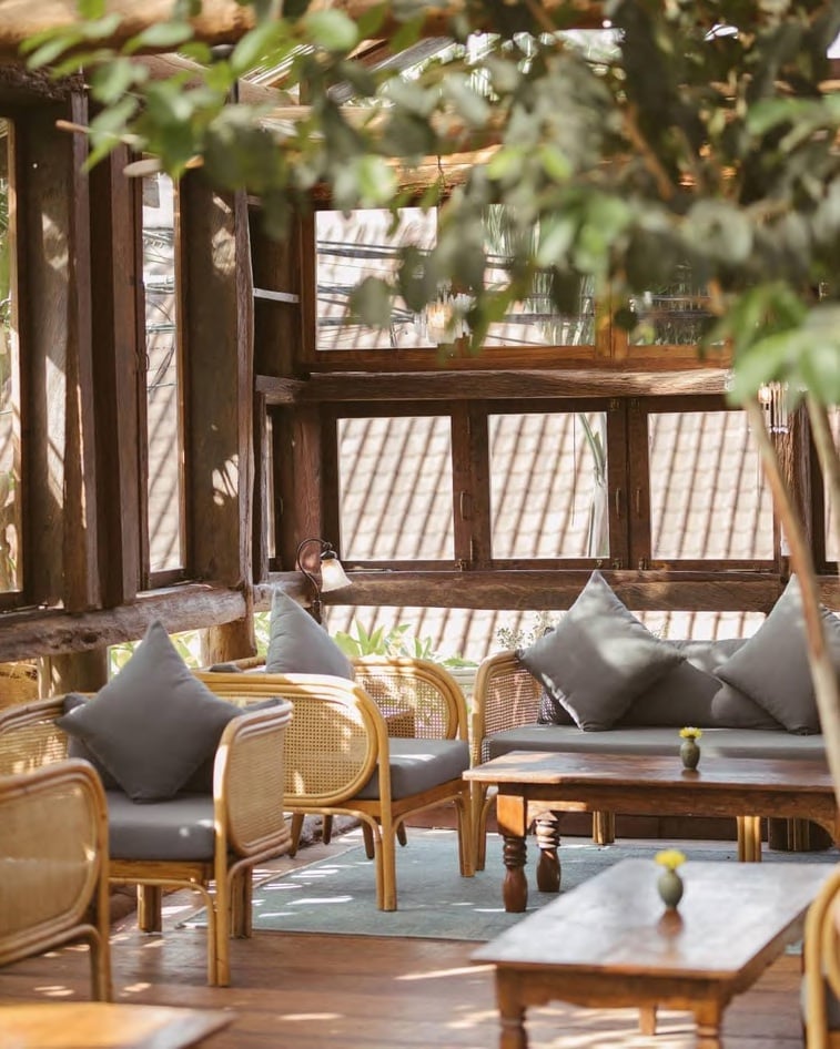Woods, Asia-Pacific — Wooden daybed lounge with grey cushions and woven rattan chairs overlooking slatted pergola, dappled sunlight across terraced deck.