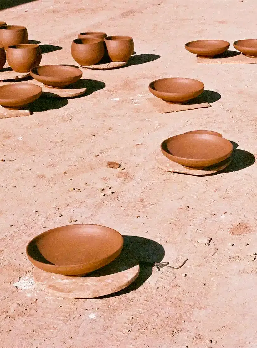 Workshops — Handmade terracotta bowls and vessels arranged on ochre earth, drying in sunlight with sharp shadows, artisanal craft workshop.