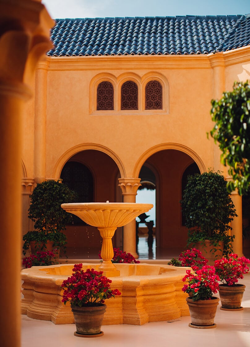 Cuixmala, Mexico — Ochre courtyard with blue-tiled roof, arched colonnades, central fountain, and potted red bougainvillea in afternoon light.