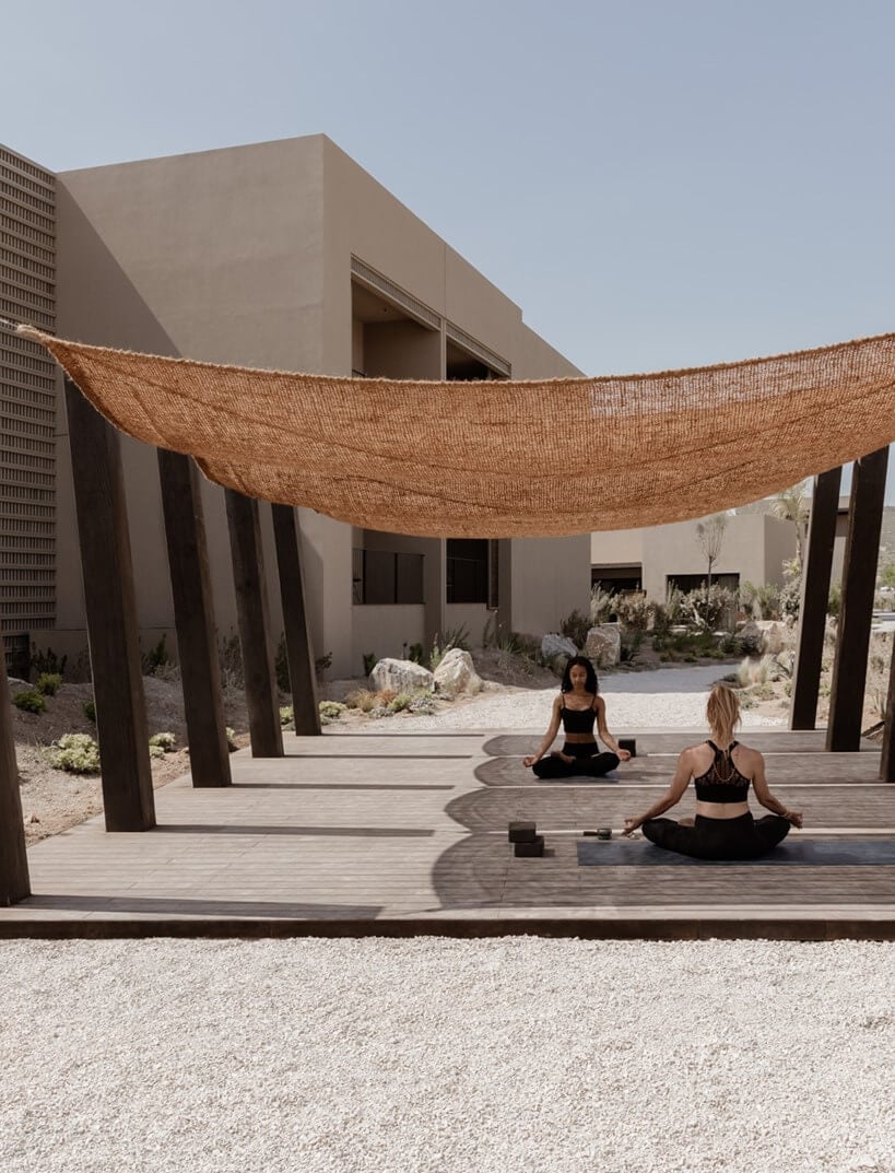 Yoga Shala, Europe — Wooden deck with woven shade canopy, minimalist architecture, two practitioners in meditation pose, desert landscaping, natural light and shadow.