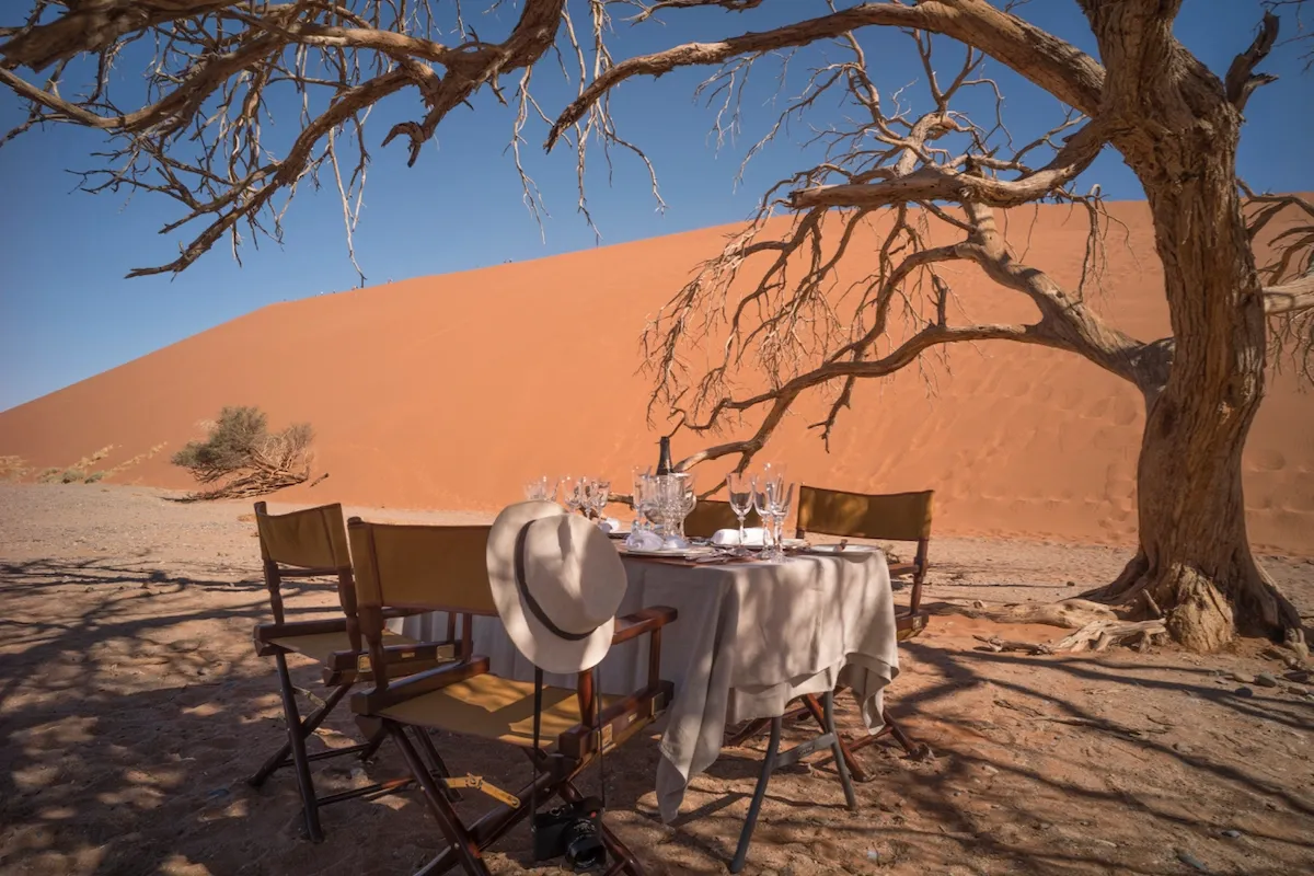 Zannier Sonop, Africa — Candlelit desert dining setup beneath dead acacia tree, white tablecloth and chairs facing red sand dune under clear sky.